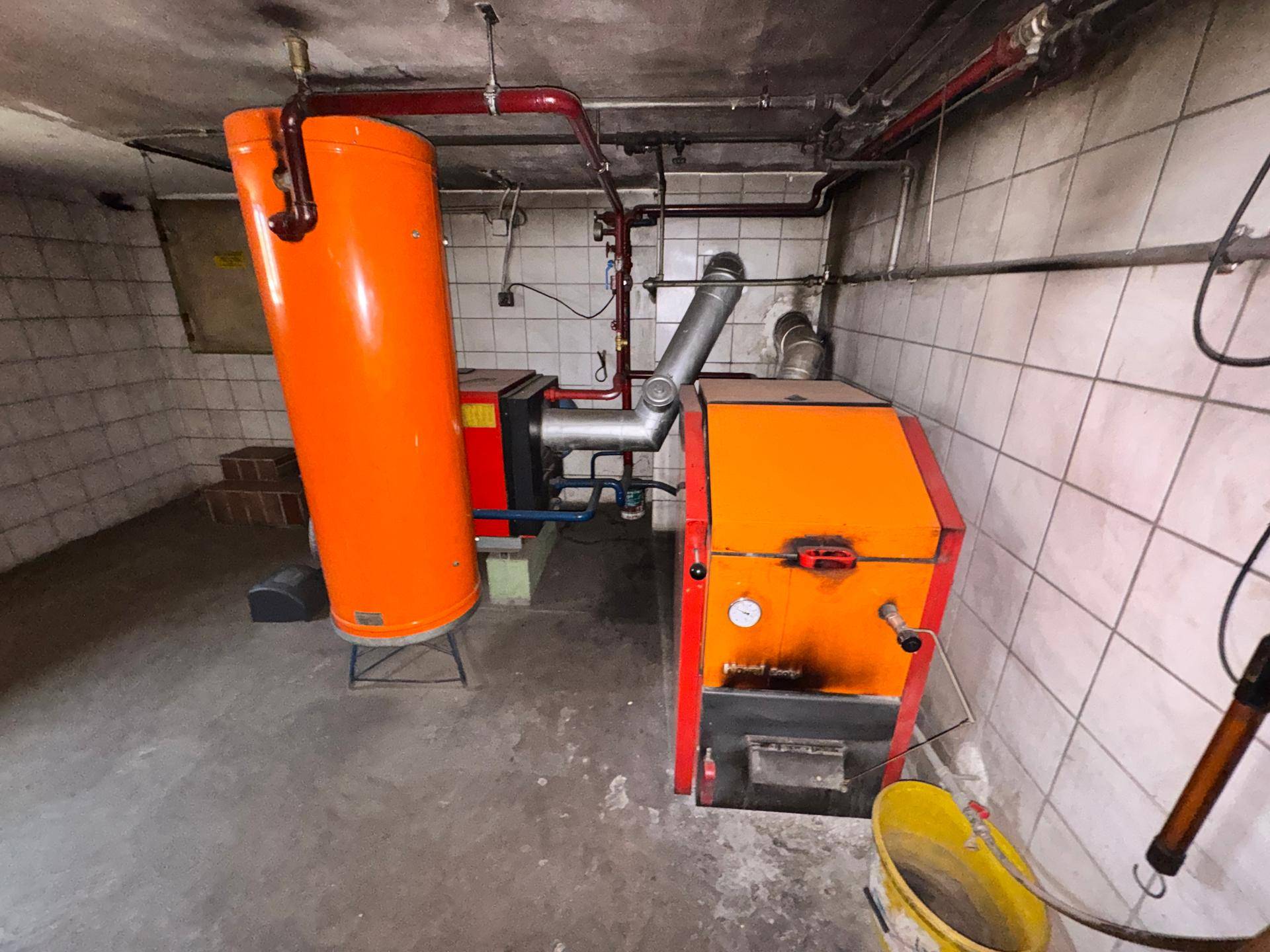 Heizungsraum im Keller mit orangefarbenem Boiler, Ofen und gefliesten Wänden.
