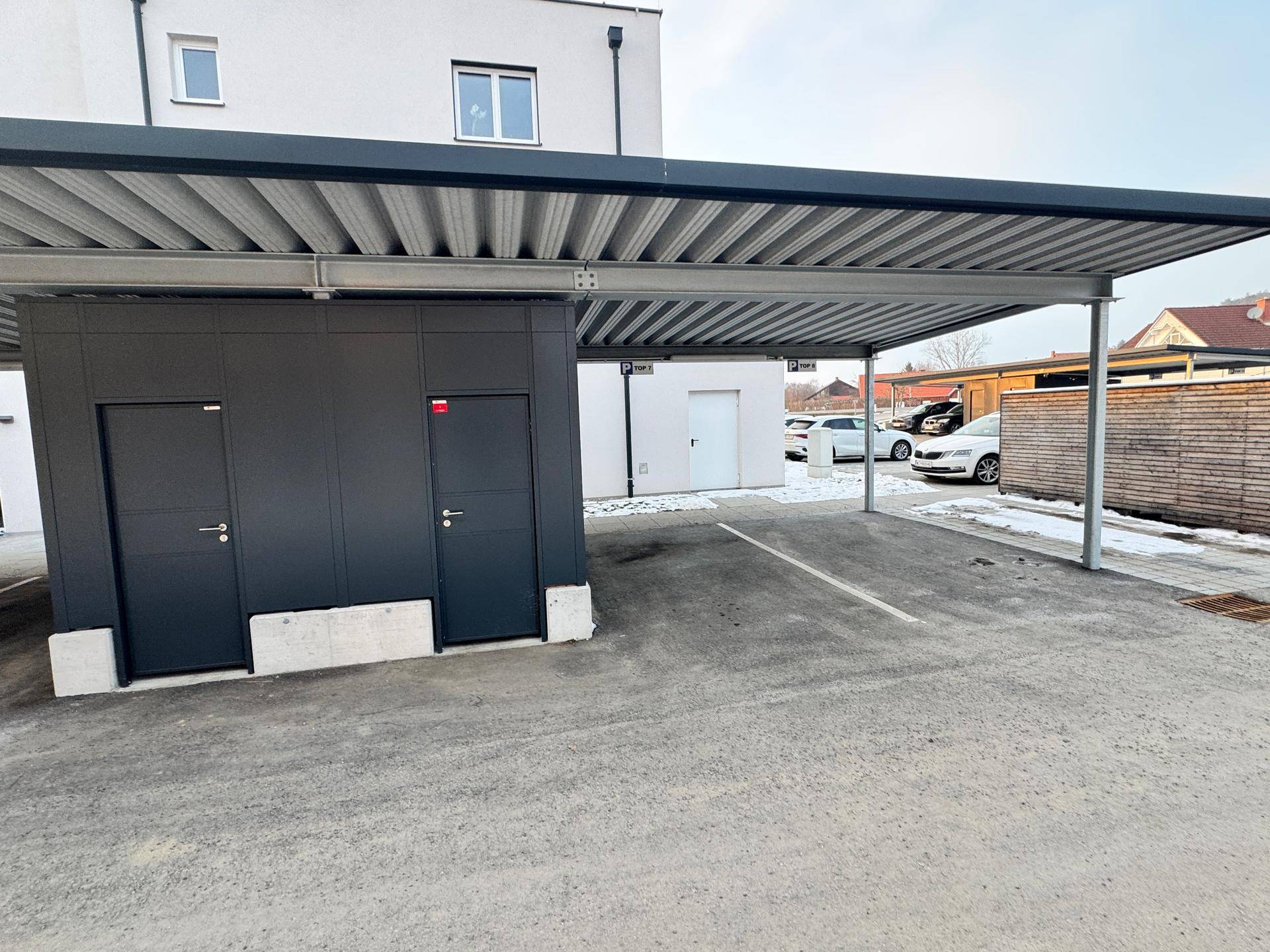 Überdachte Parkplätze (Carports) neben dem Gebäude mit Abstellräumen.