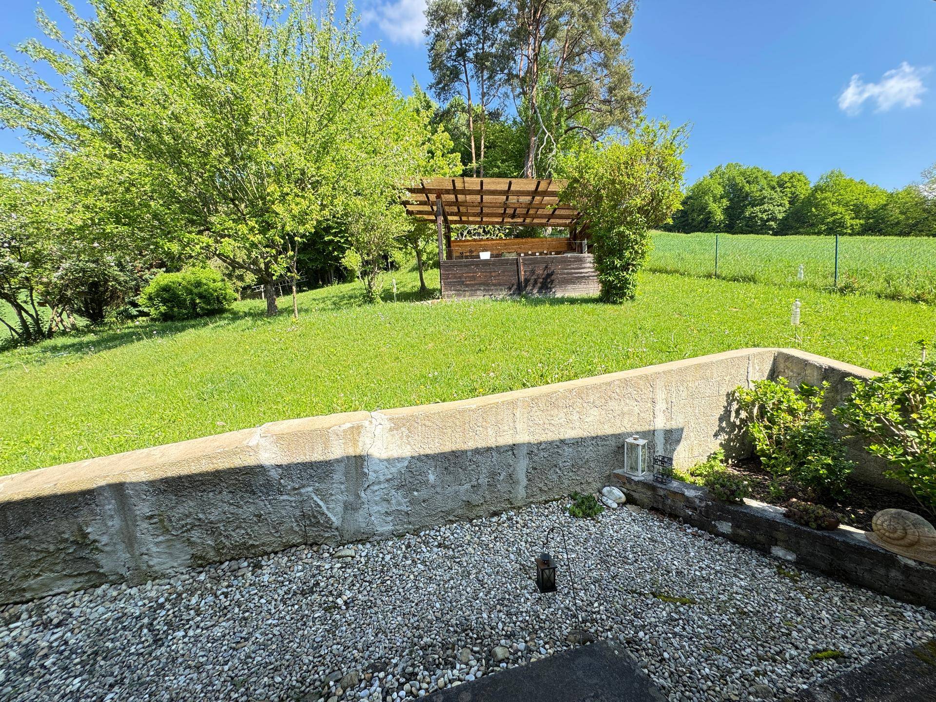 Kiesfläche im Garten mit einer überdachten Holzpergola und Blick ins Grüne.