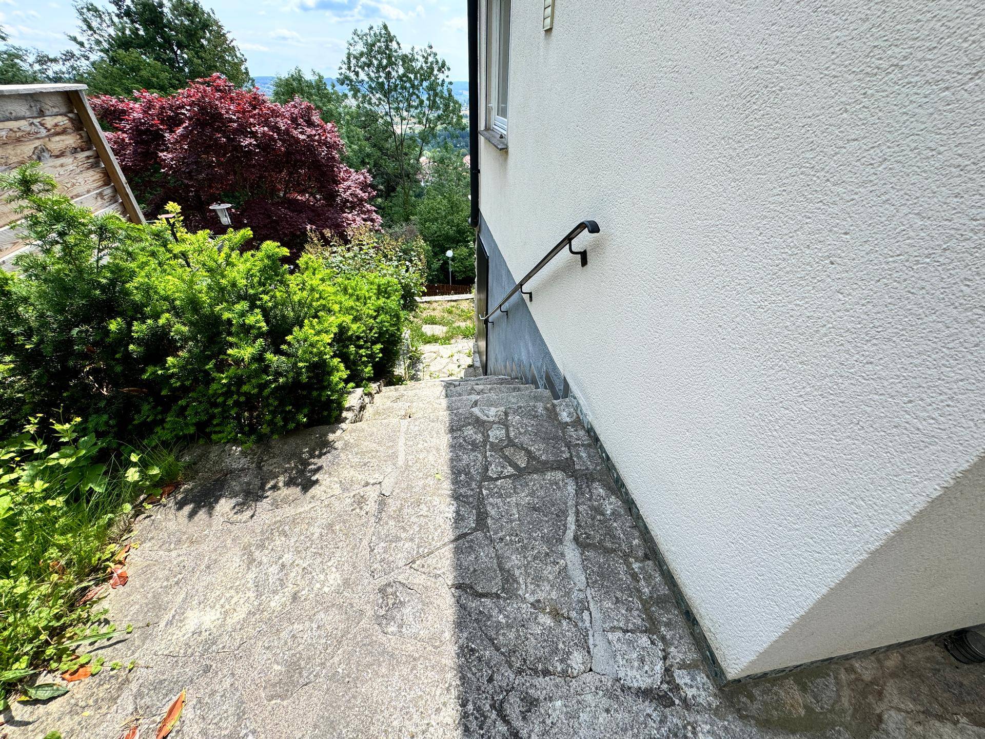 Steinstufen führen entlang einer hellen Hauswand hinunter, umgeben von grünen Büschen.