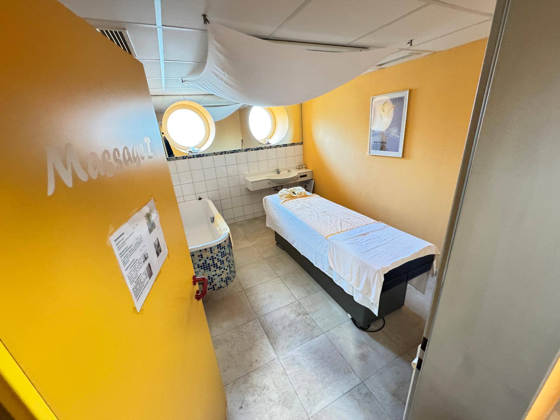 Massageraum mit Liege, Badewanne und runden Fenstern.