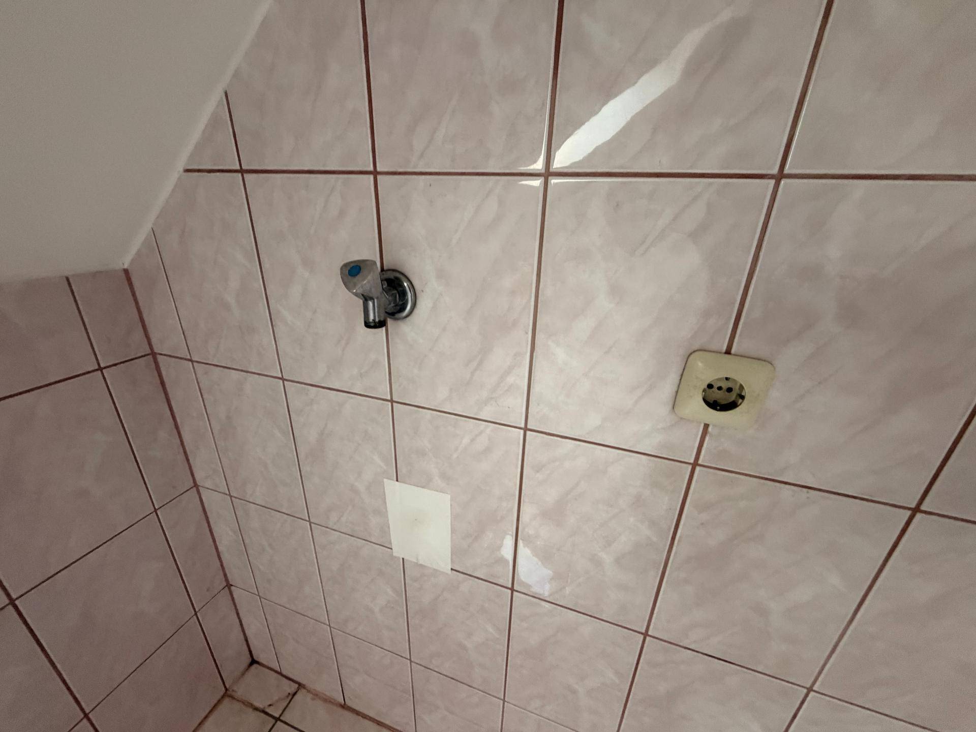 Detailansicht der rosa gefliesten Wand im Badezimmer mit Steckdose und Wasseranschluss.