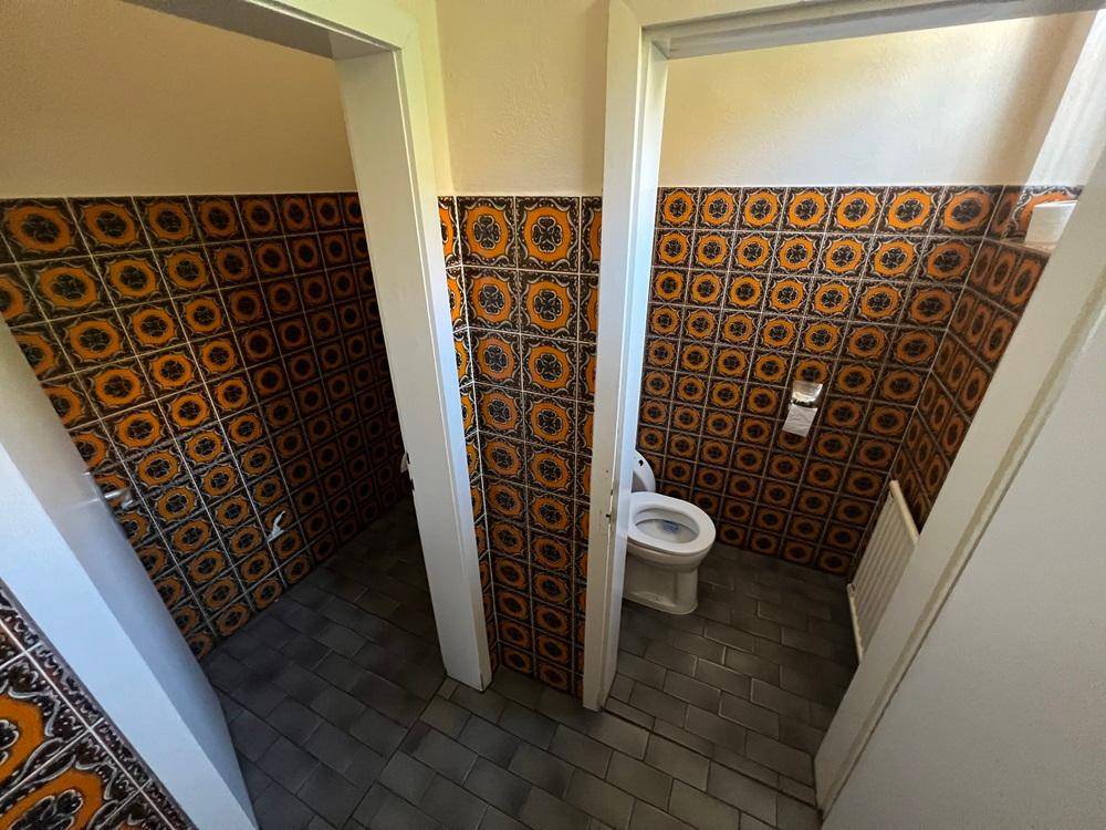 Toilettenbereich mit orange-gemusterten Wandfliesen und grauen Bodenfliesen, getrennt durch Türen.