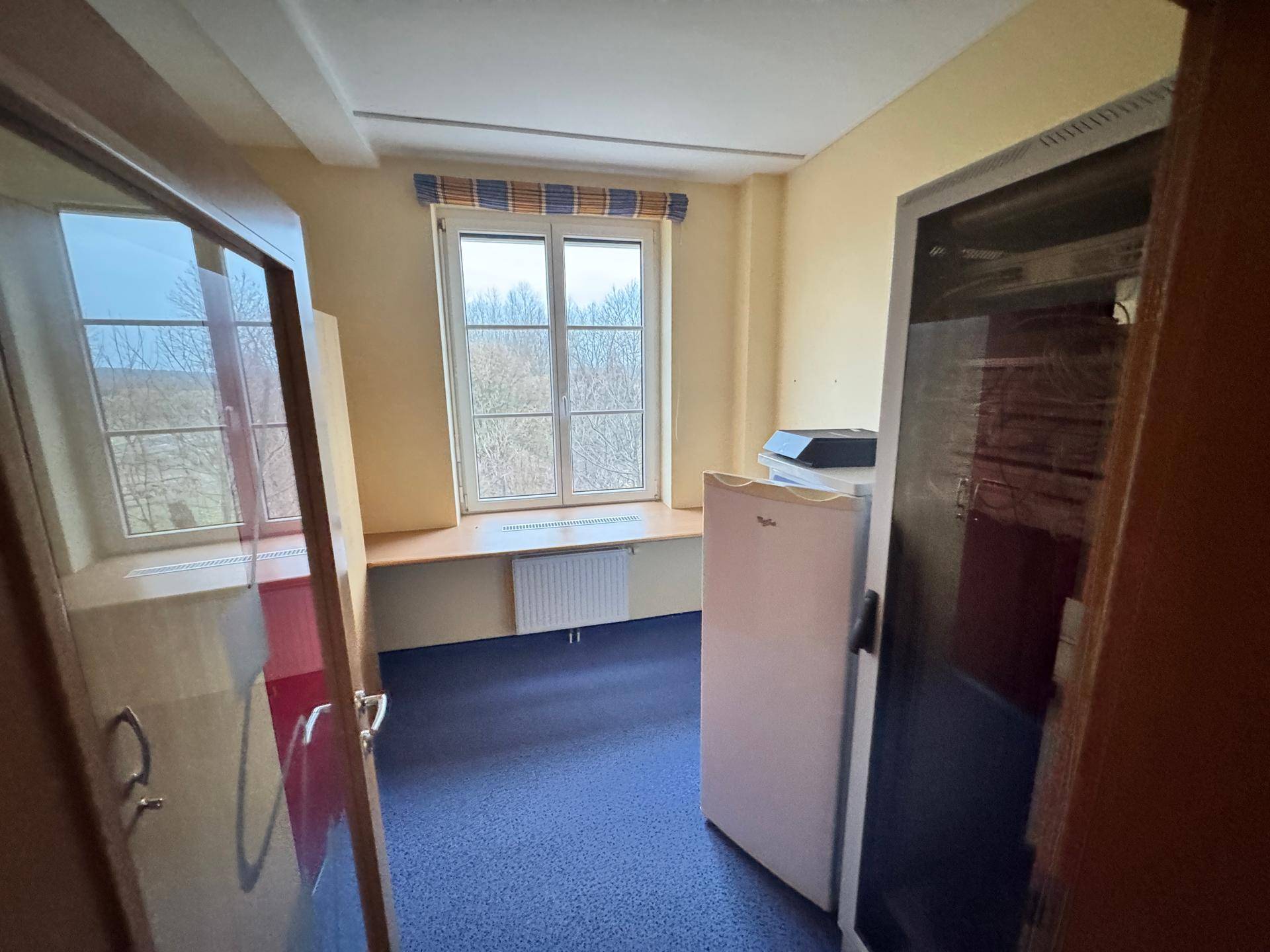 Kleiner Büroraum mit Fenster, blauem Teppichboden und einem Kühlschrank.