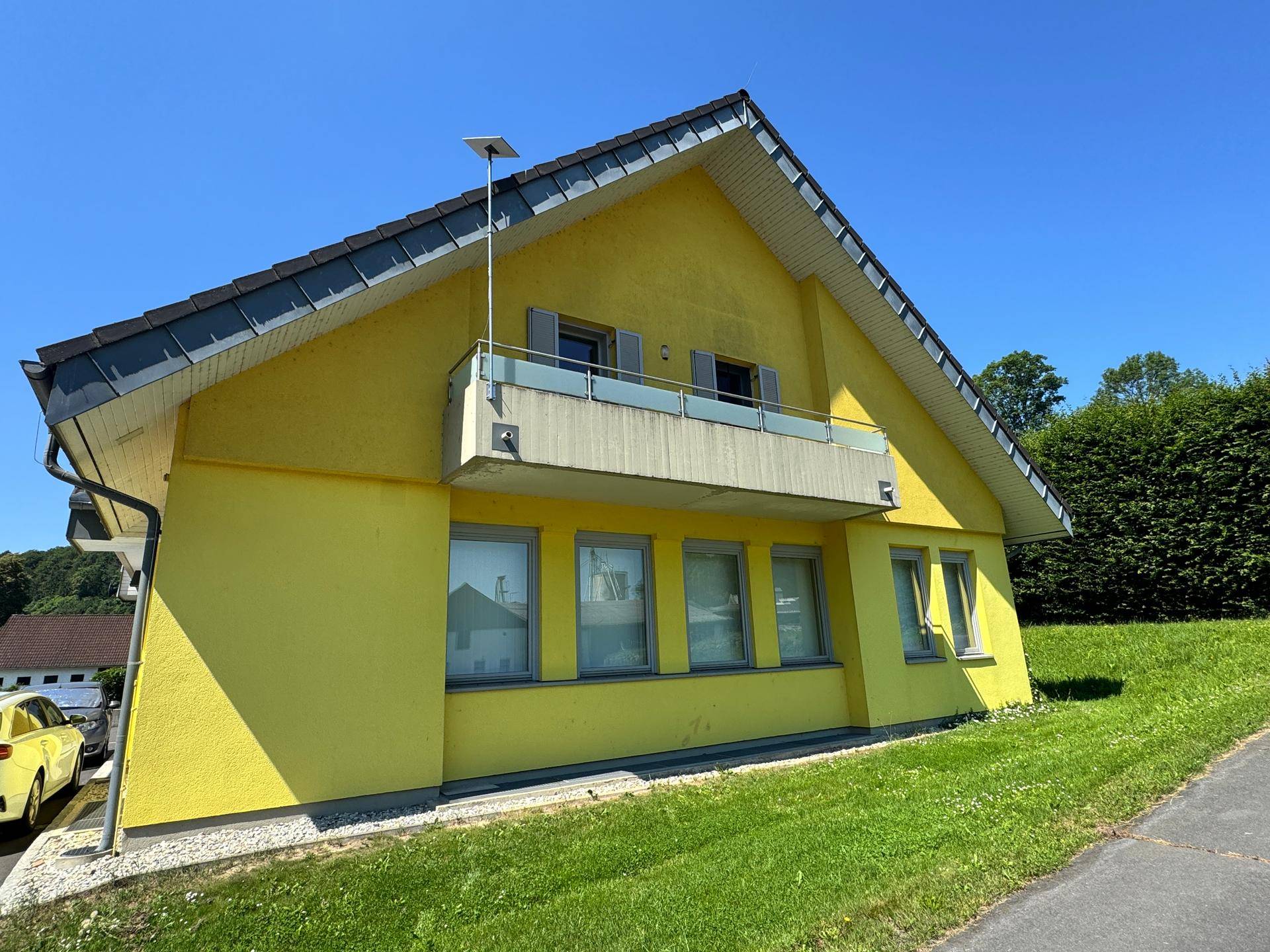 Seitenansicht eines gelben Hauses mit Satteldach, kleinem Balkon und grüner Umgebung unter blauem Himmel.