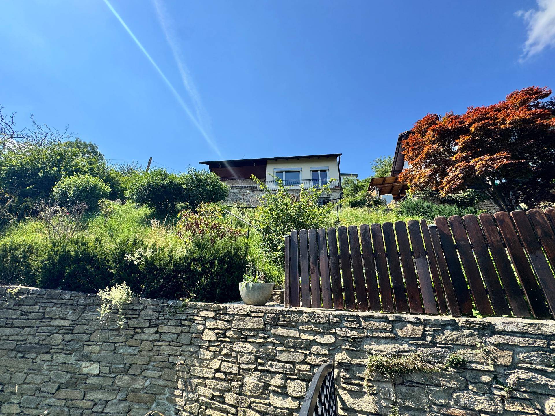 Haus auf einem Hügel mit Steinfassade, Holzzaun und üppiger Vegetation unter blauem Himmel.