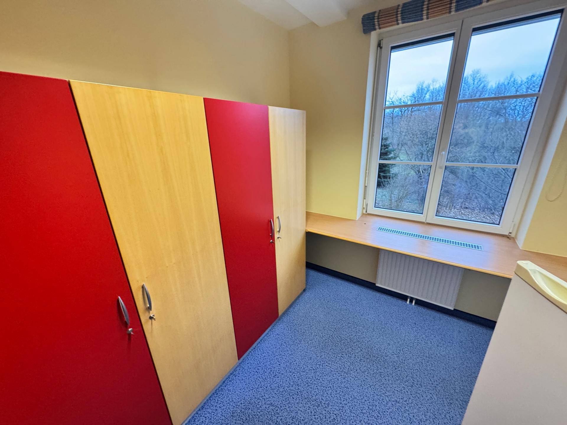 Kleiner Büroraum mit bunten Schränken, blauem Teppichboden und einem Fenster.