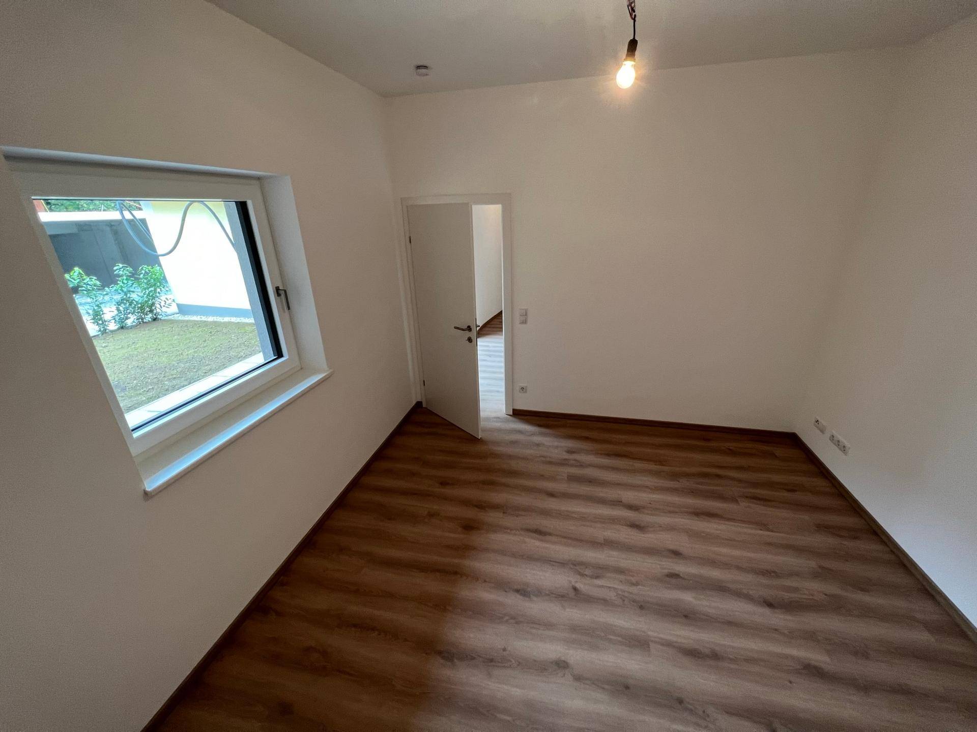 Leeres Zimmer mit Laminatboden und einem Fenster, das den Blick ins Grüne freigibt.