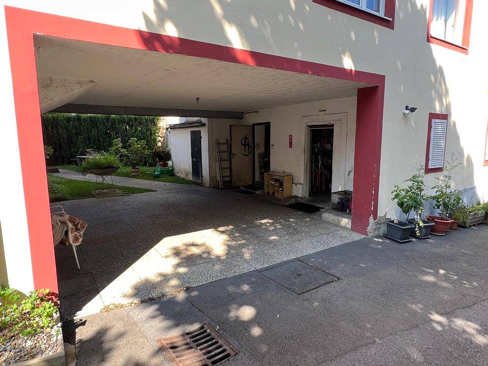 Überdachter Parkbereich neben dem Gebäude mit Zugang zu Lagerräumen und Garten.