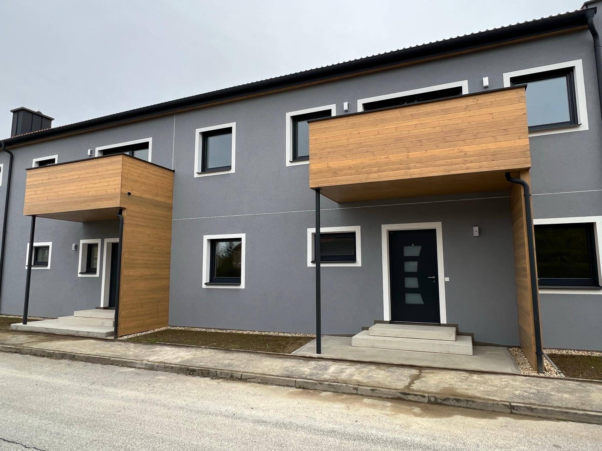 Fassade eines modernen Wohngebäudes mit grauer Putzfassade, Holzelementen und dunklen Eingangstüren.
