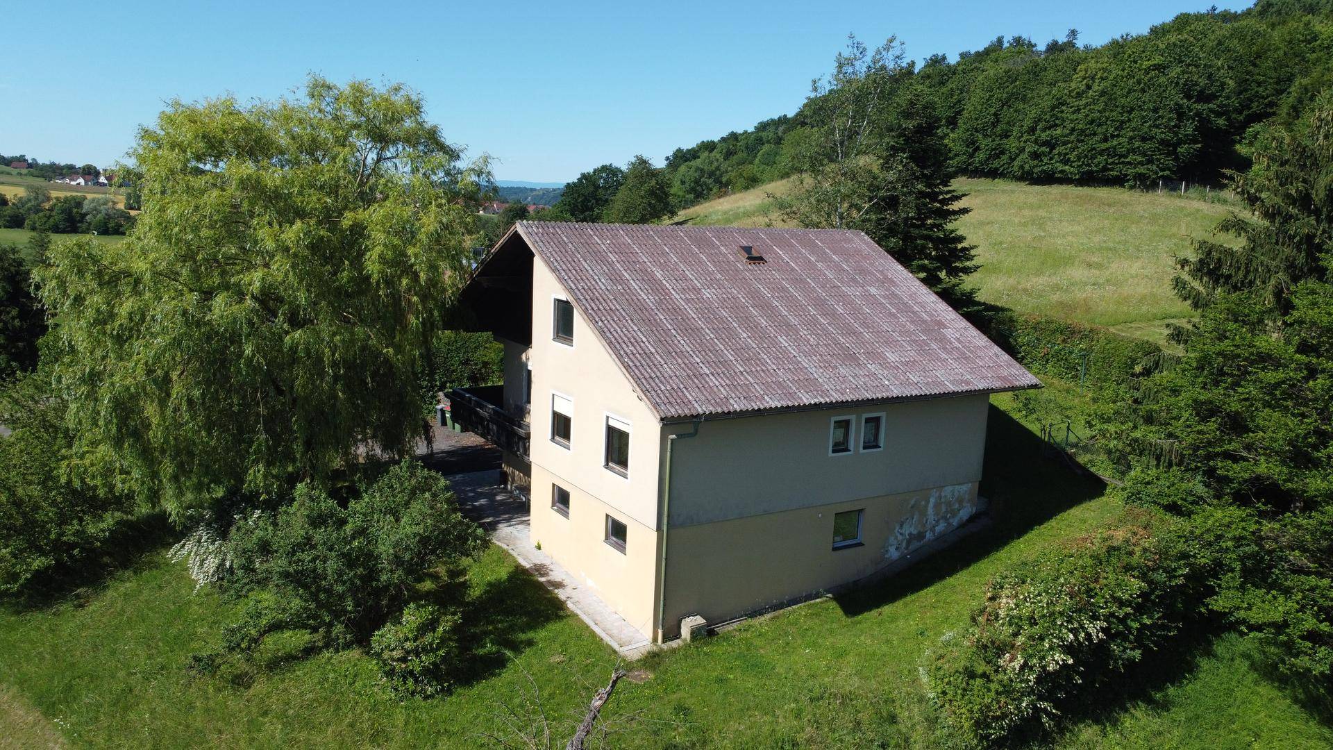 Luftaufnahme des Hauses mit Satteldach, umgeben von grünen Hügeln und Bäumen unter blauem Himmel.
