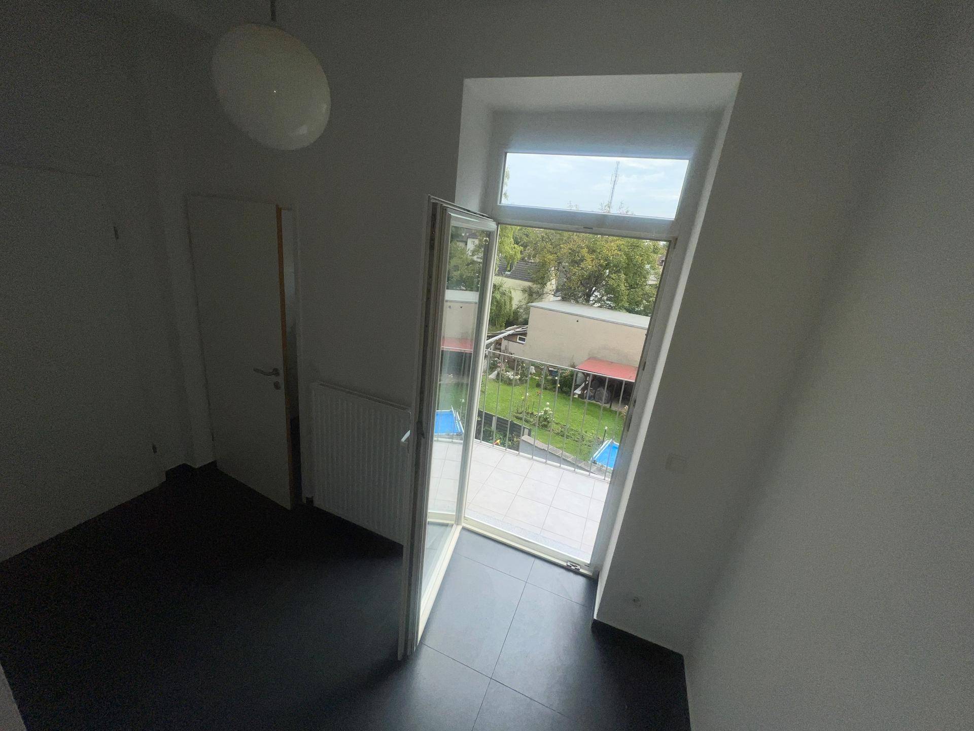 Kleiner Raum mit dunklem Fliesenboden und Zugang zu einem Balkon mit Blick ins Grüne.