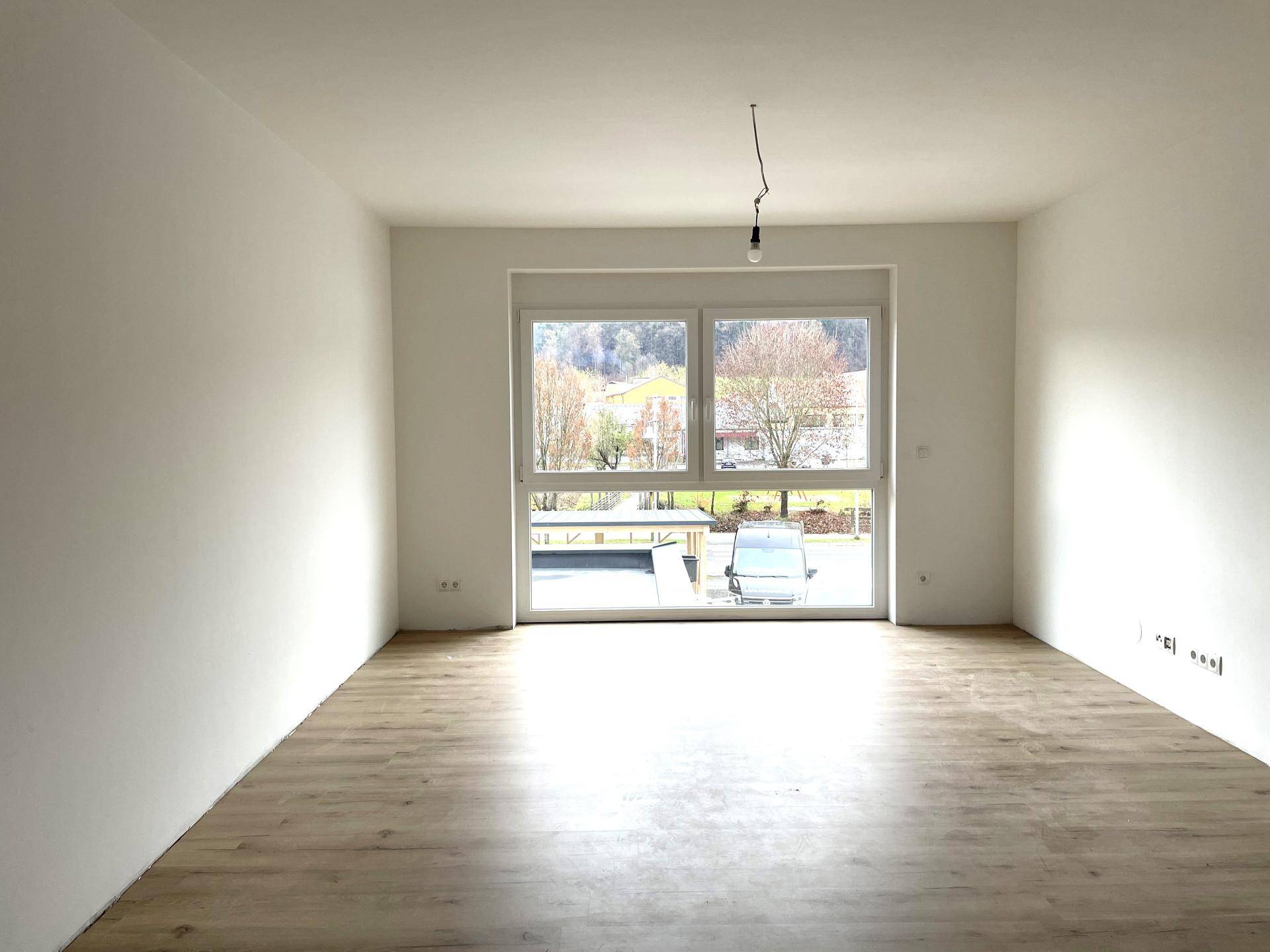 Heller Wohnraum mit großem Fenster, das einen Blick ins Grüne bietet, und Laminatboden.