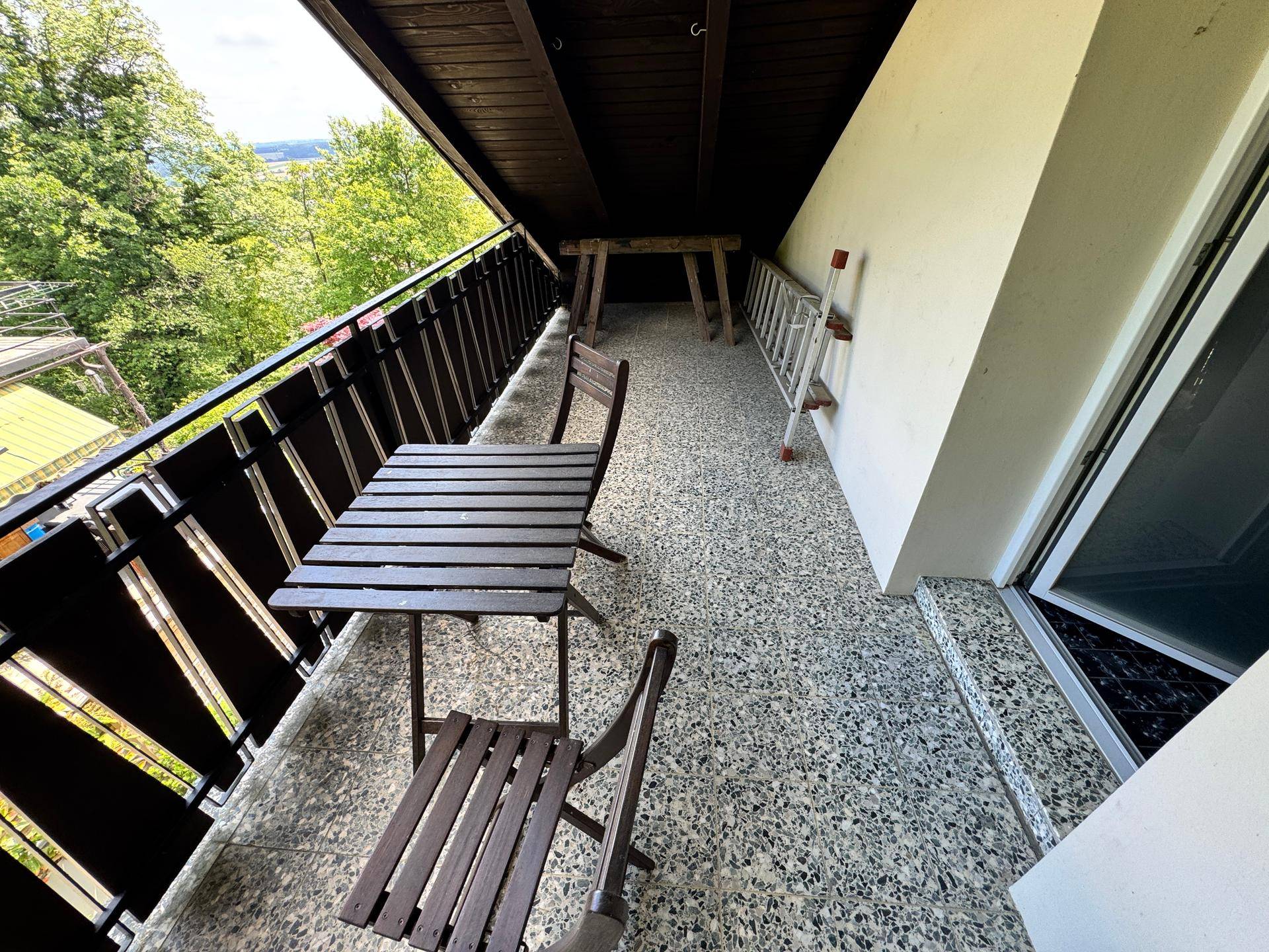 Schmaler Balkon mit kleinem Holztisch und Stühlen, bietet einen Blick auf die umliegenden Bäume.