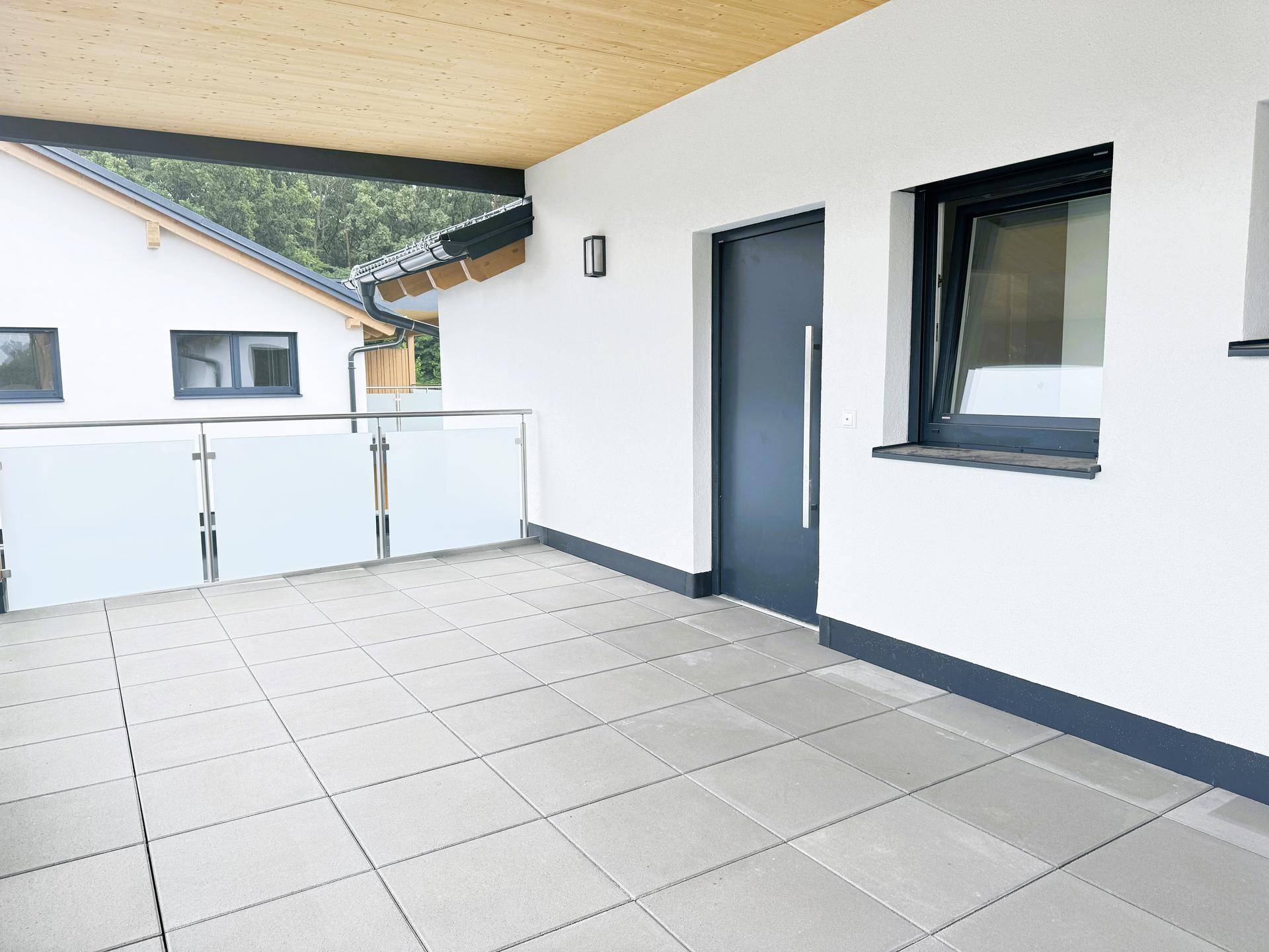 Überdachte Terrasse mit Fliesenboden und Zugang zum Gebäude in einem Neubau.