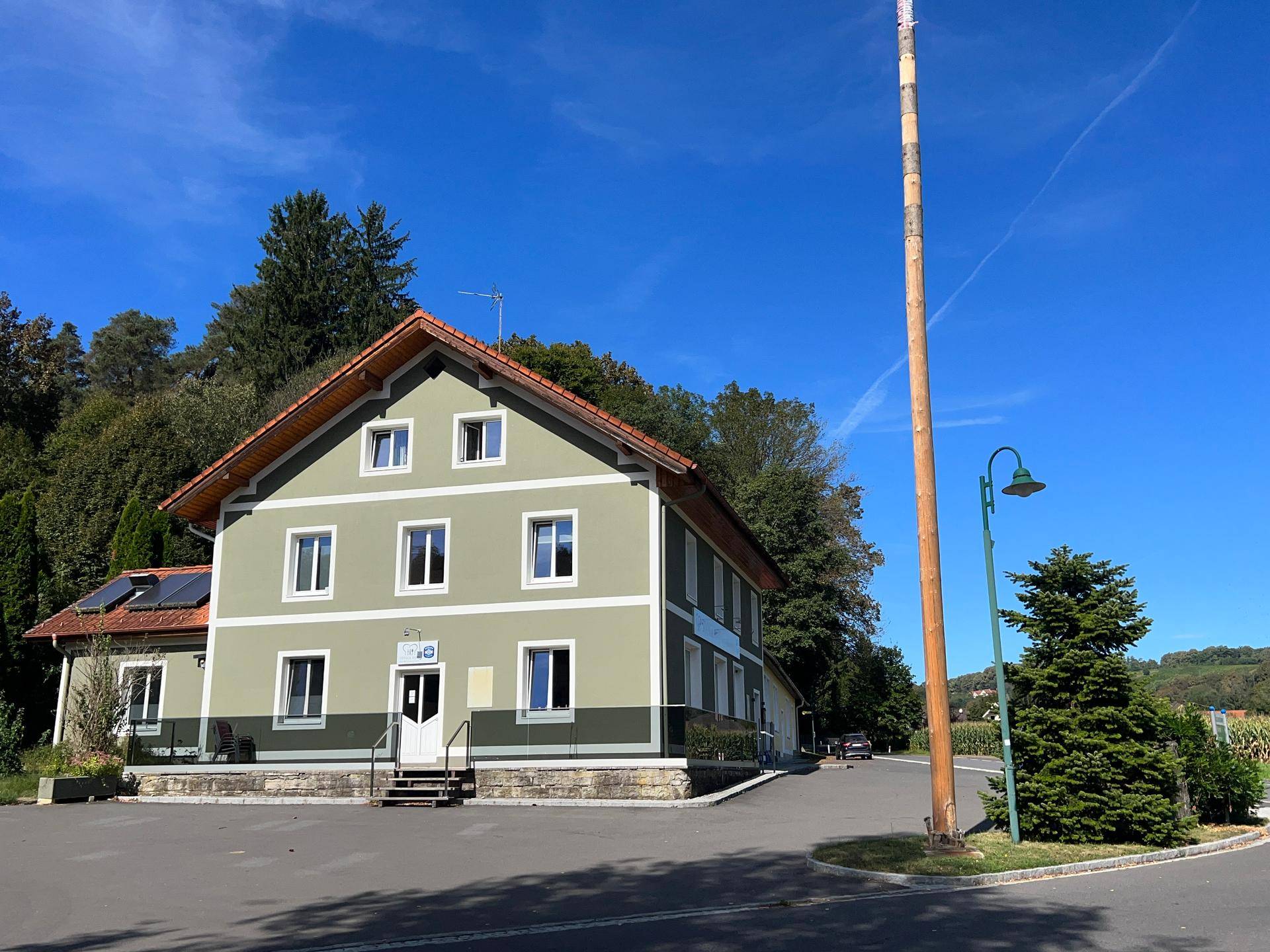 Gebäudeansicht mit großem Parkplatz, Maibaum und angrenzender Straße, umgeben von grüner Natur.