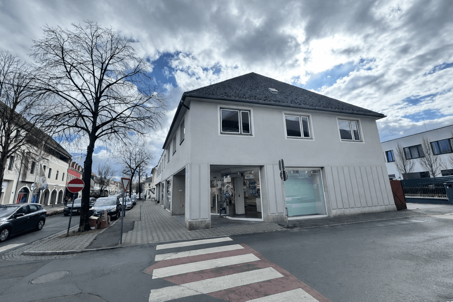 Gebäude von der Straße aus gesehen, mit einem Zebrastreifen im Vordergrund und parkenden Autos.