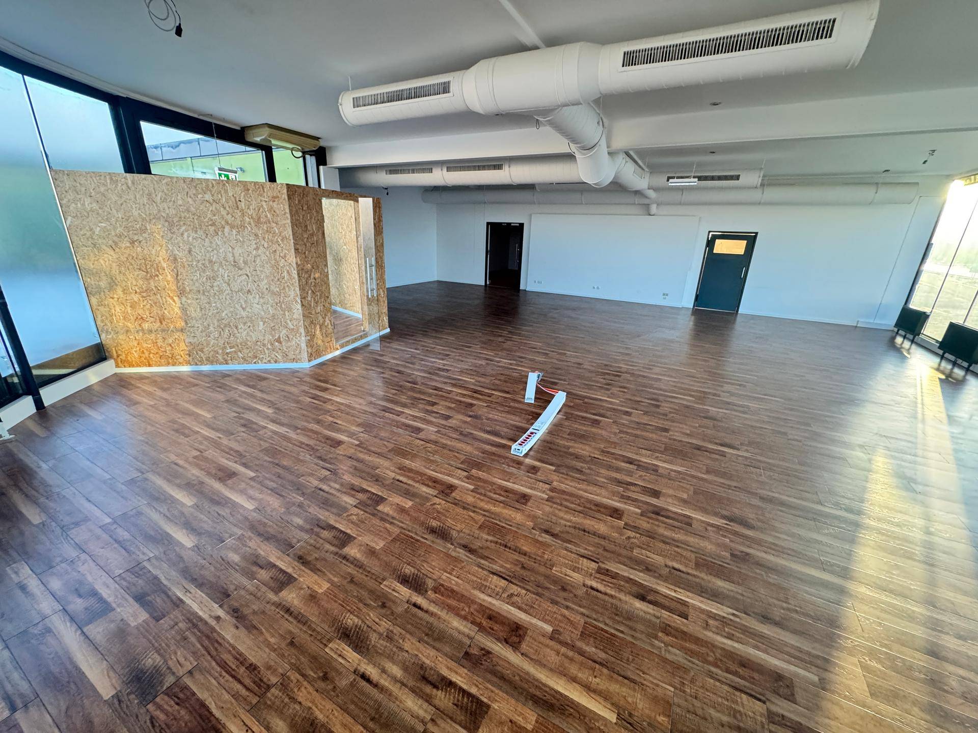 Weitläufiger Innenraum mit Holzboden, freiliegenden Rohren an der Decke und viel Tageslicht.