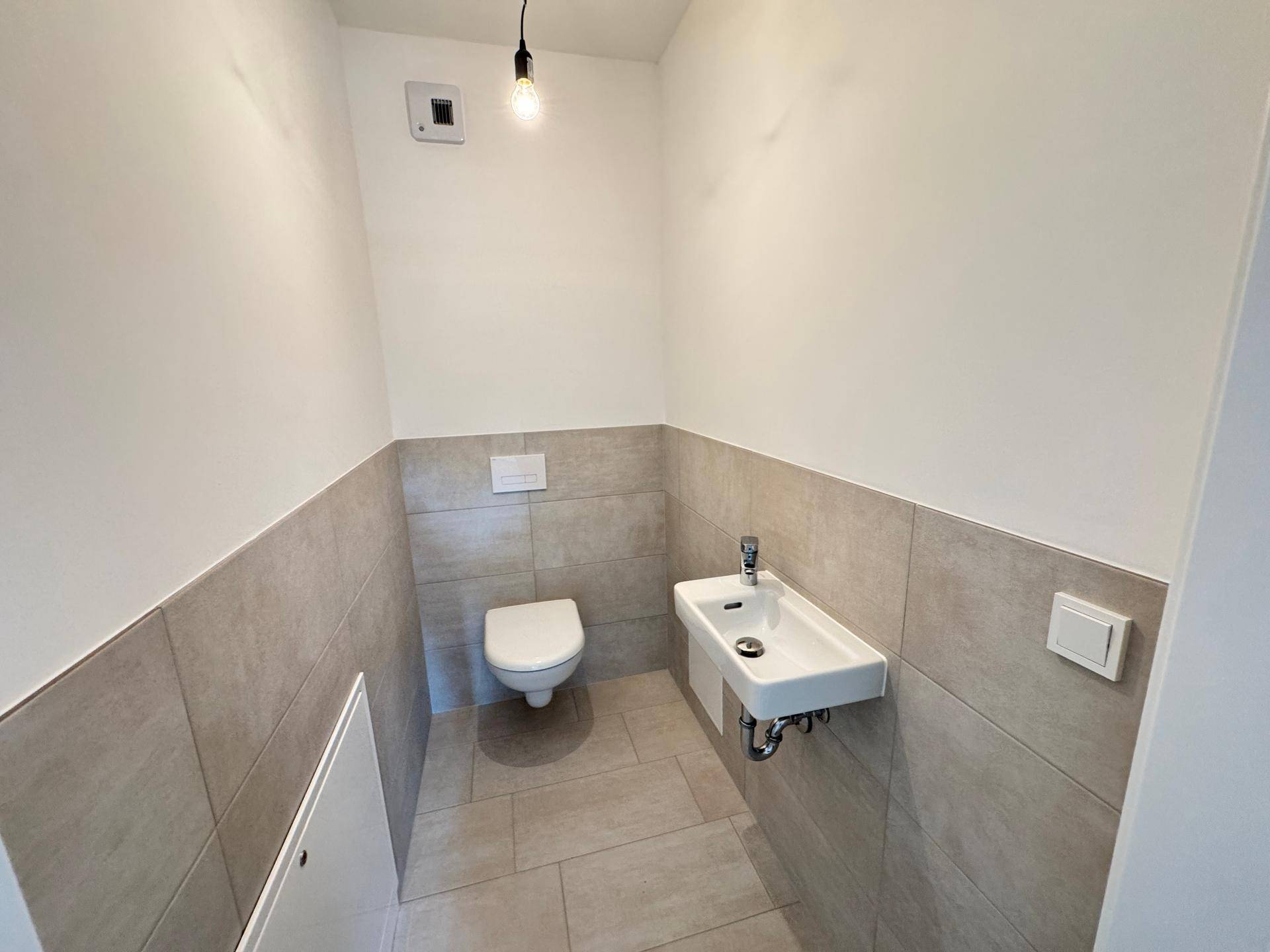 Separate Gästetoilette mit Wand-WC, kleinem Waschbecken und hellen Fliesen.