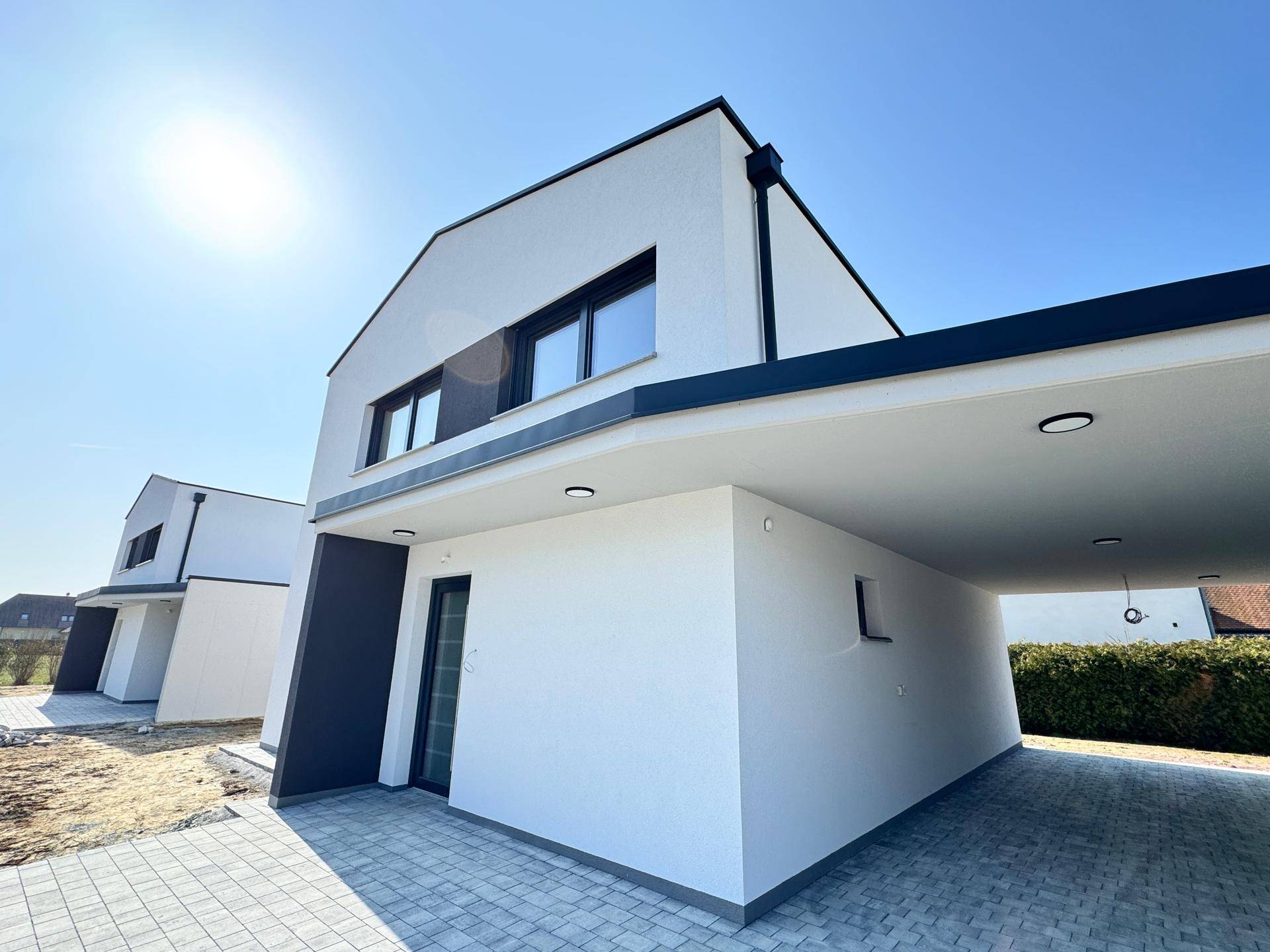 Modernes Einfamilienhaus mit weißer Fassade, schwarzem Dach und integriertem Carport unter blauem Himmel.