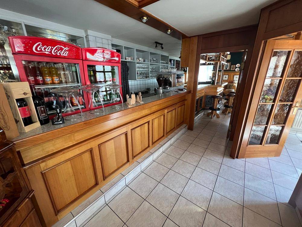 Bar- und Gastbereich mit Holzeinrichtung, Fliesenboden und einer Theke mit Gläsern und Kaffeemaschine.