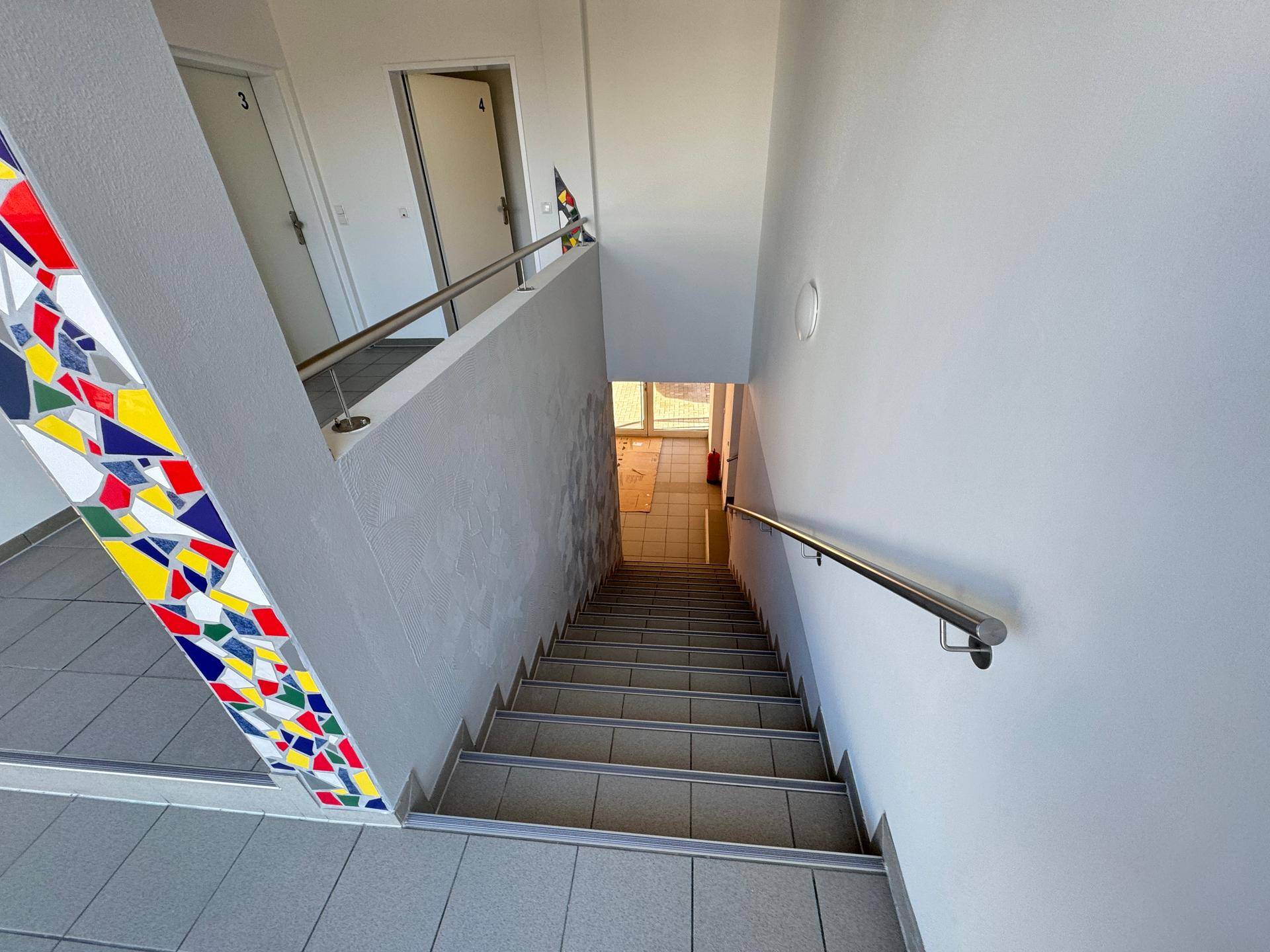 Treppenhaus mit farbiger Mosaikverzierung an der Wand und Fliesenboden.