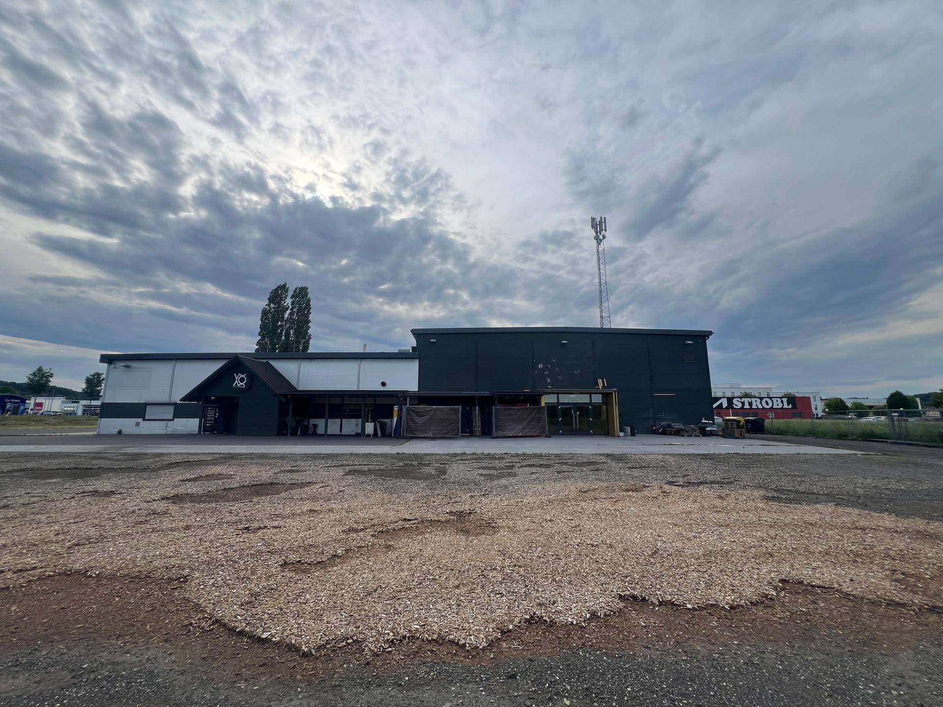 Gewerbegebäude mit Schotterparkplatz und weiteren Gebäuden im Hintergrund unter bewölktem Himmel.