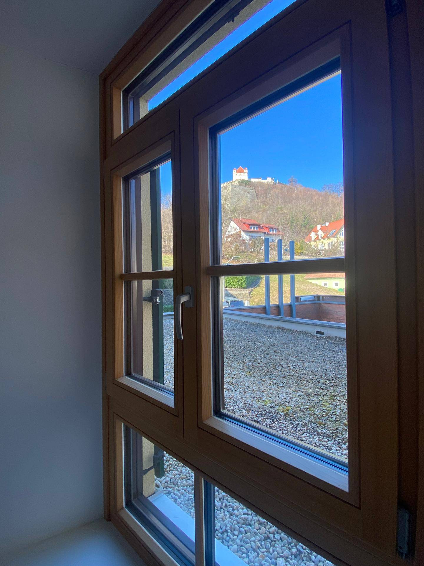 Ausblick aus einem Holzfenster auf einen Hügel mit Burg und umliegende Häuser.