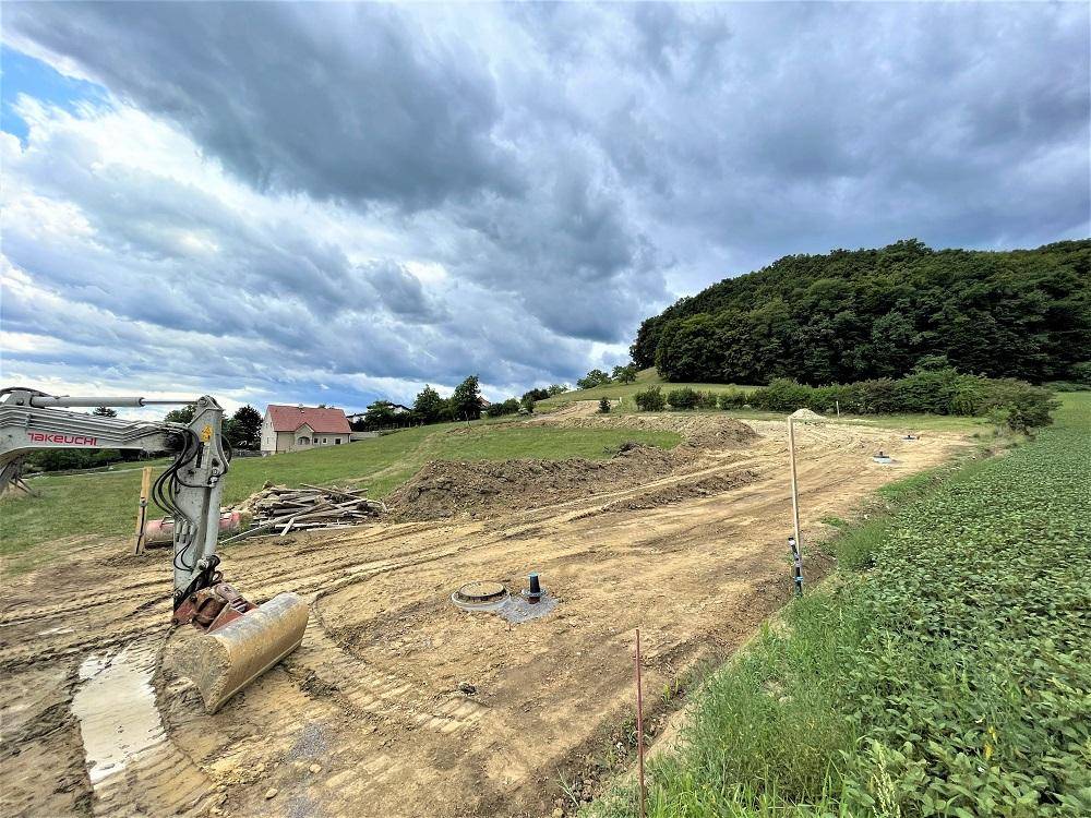 Baustelle mit Bagger und vorbereitetem Gelände vor bewaldeten Hügeln unter bewölktem Himmel.
