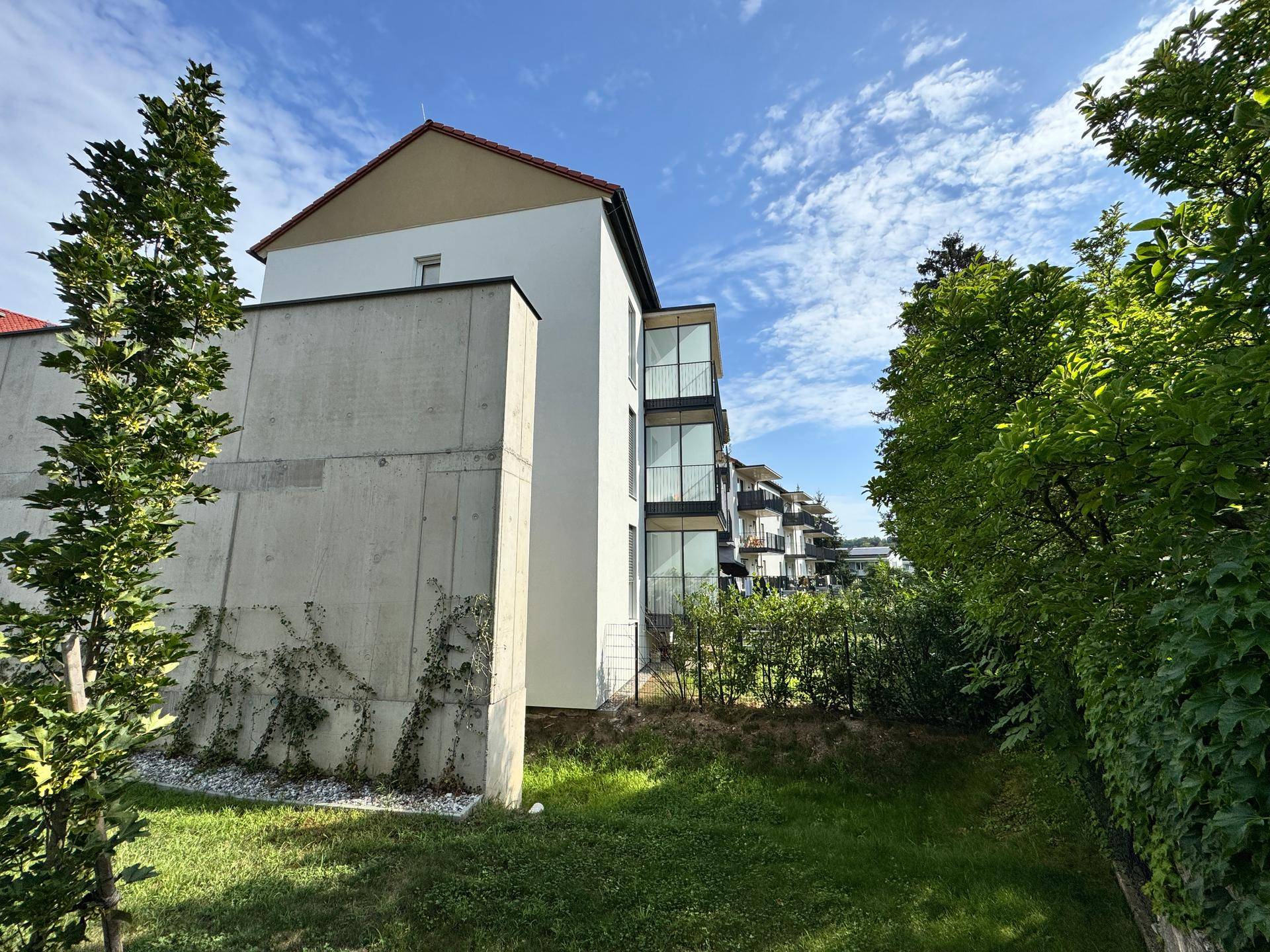 Moderne Gebäudefassade mit Betonwänden und grüner Bepflanzung in sonniger Umgebung.