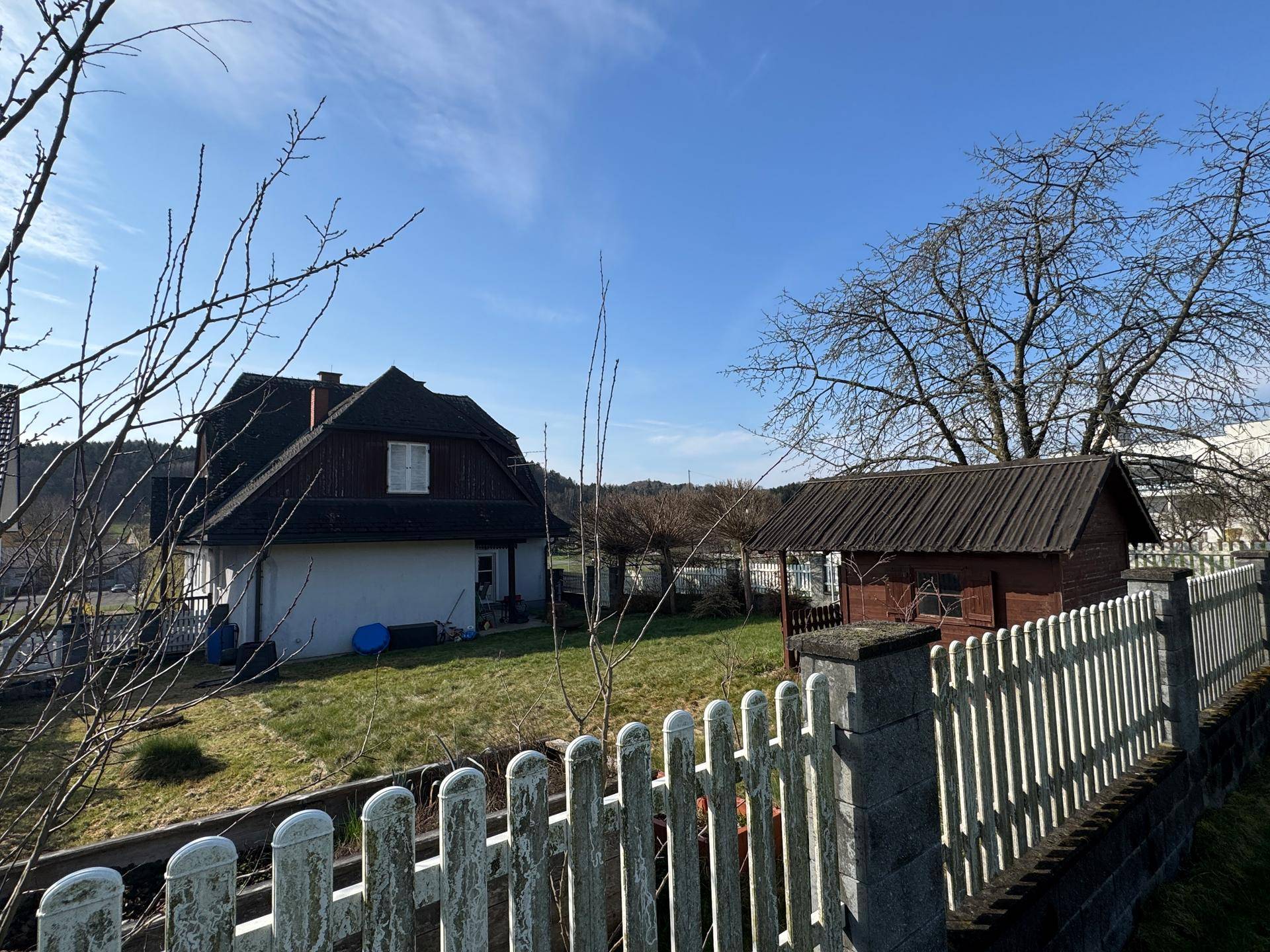 Blick auf das Haus und den Holzschuppen von der Gartenseite, umgeben von einem Zaun.