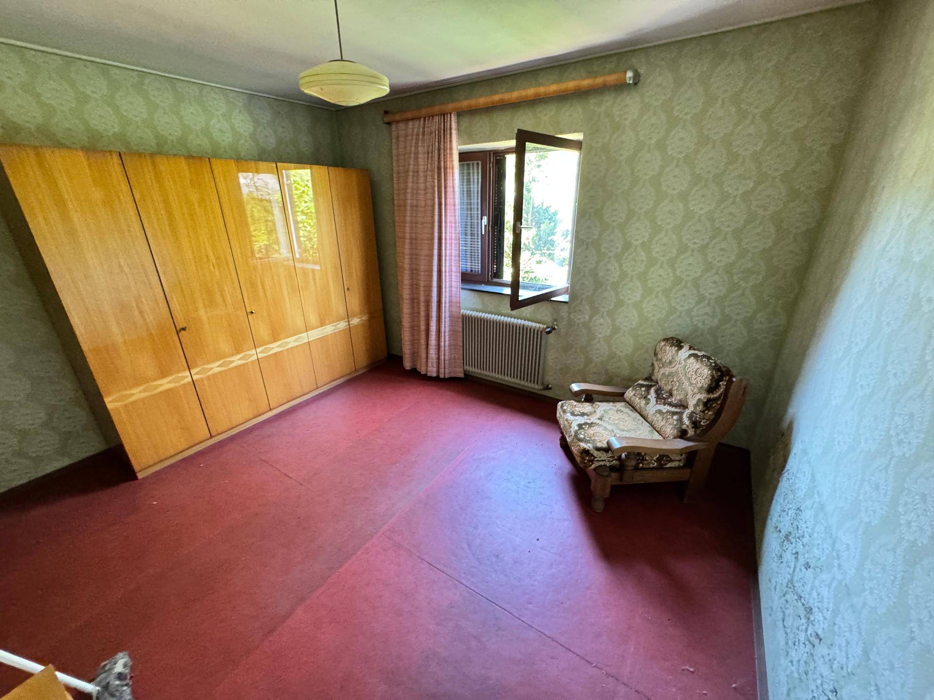 Schlafzimmer mit grüner Mustertapete, Holzschrank und einem Sessel, roter Bodenbelag.