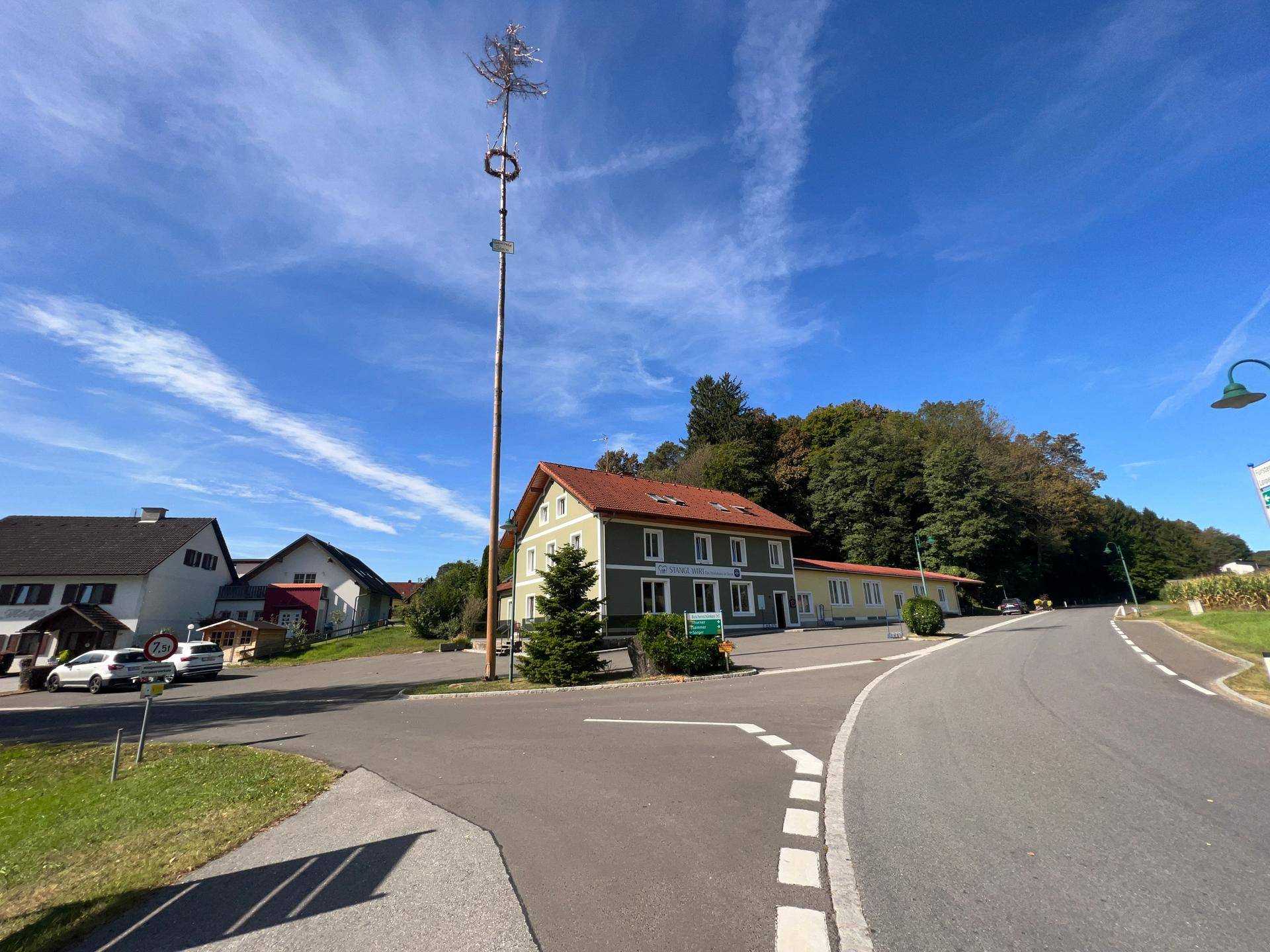Panoramablick auf das Gebäude mit Maibaum, umgeben von ländlicher Umgebung und einer Straße.