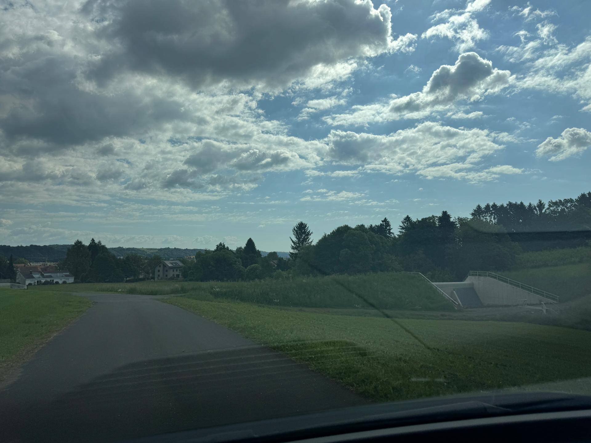 Straße führt durch eine grüne Landschaft mit Häusern in der Ferne und einer Betonkonstruktion.