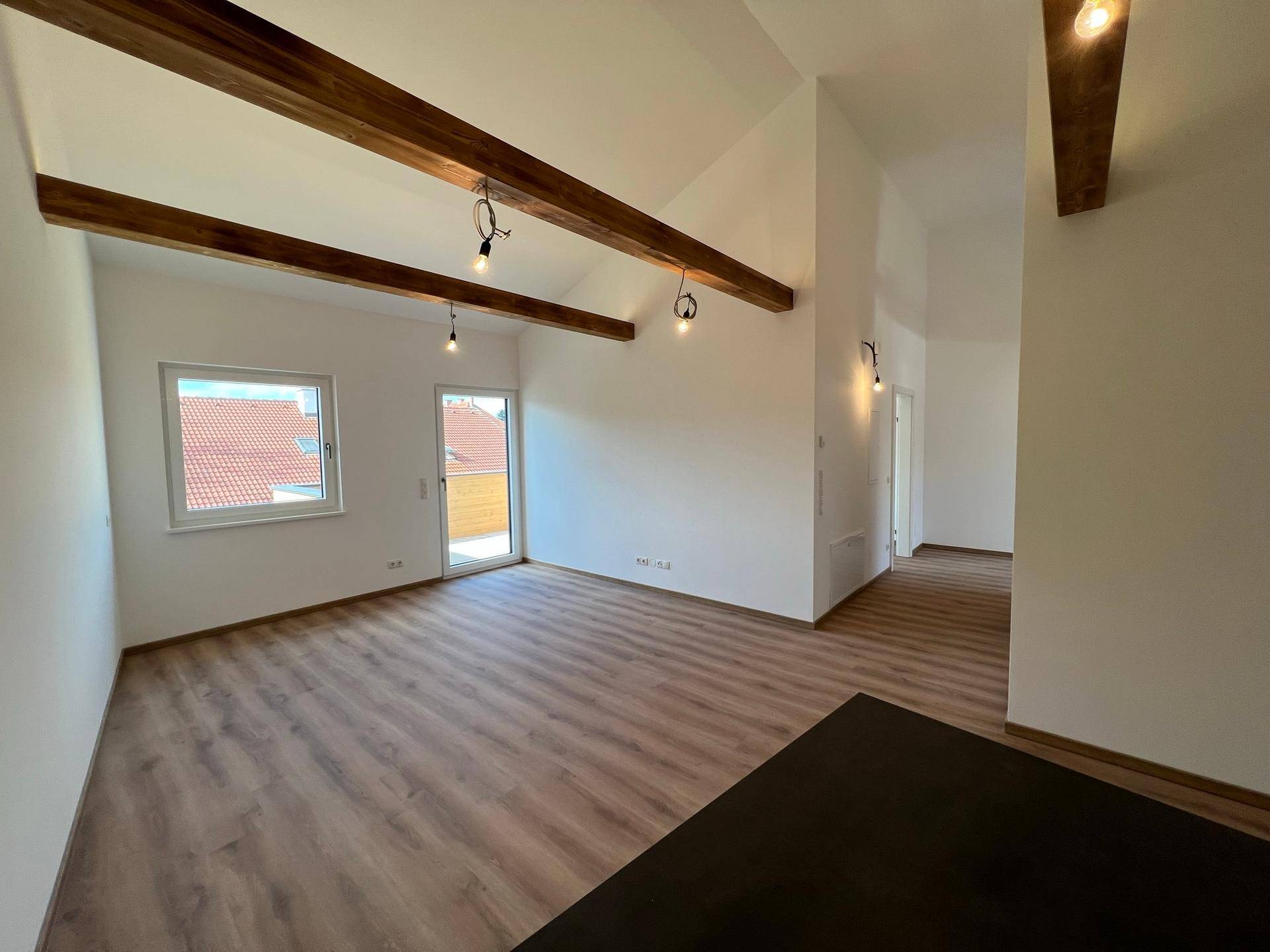 Geräumiger Wohnbereich mit hohen Decken, sichtbaren Holzbalken und hellem Laminatboden.