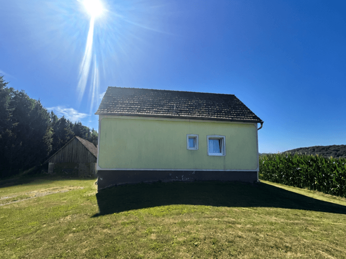 Gelbes Haus mit dunklem Dach und einem rustikalen Holzgebäude im Hintergrund.