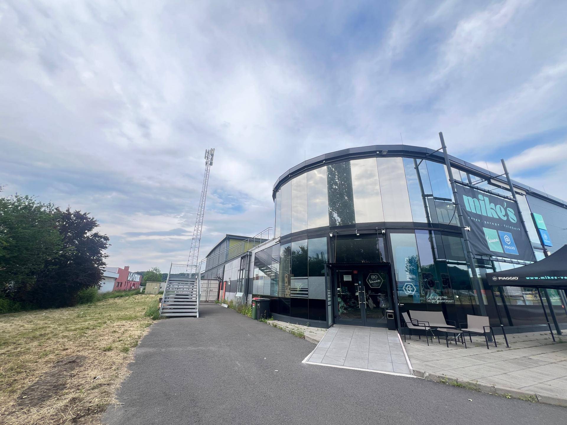 Ansicht des modernen Gebäudes mit runder Glasfront und Eingangsbereich unter leicht bewölktem Himmel.