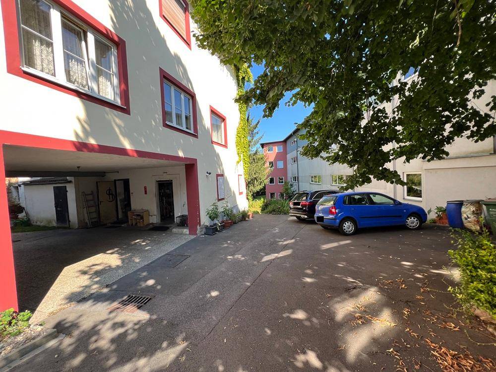 Außenbereich mit Parkplätzen und Zufahrt neben dem Gebäude, umgeben von Bäumen und Grünflächen.
