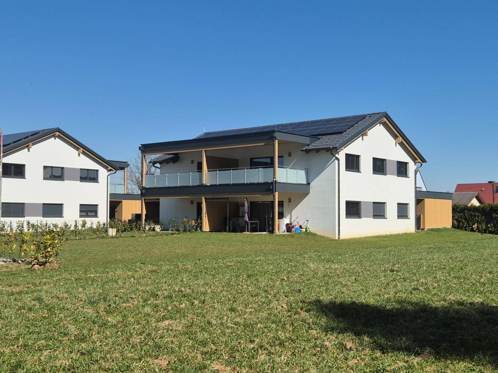 Modernes Wohnhaus mit Balkon, ebenerdiger Terrasse und großzügigem Rasenbereich unter blauem Himmel.