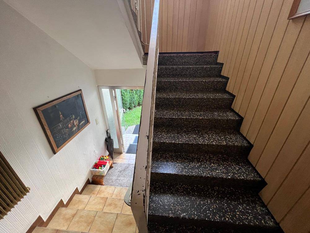 Treppenhaus mit dunklen Terrazzo-Stufen, Holzvertäfelung und Blick auf den Eingangsbereich.