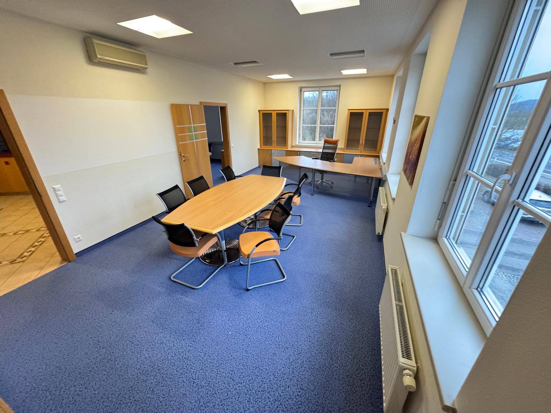 Geräumiger Büroraum mit blauem Teppichboden, Konferenztisch und großen Fenstern.