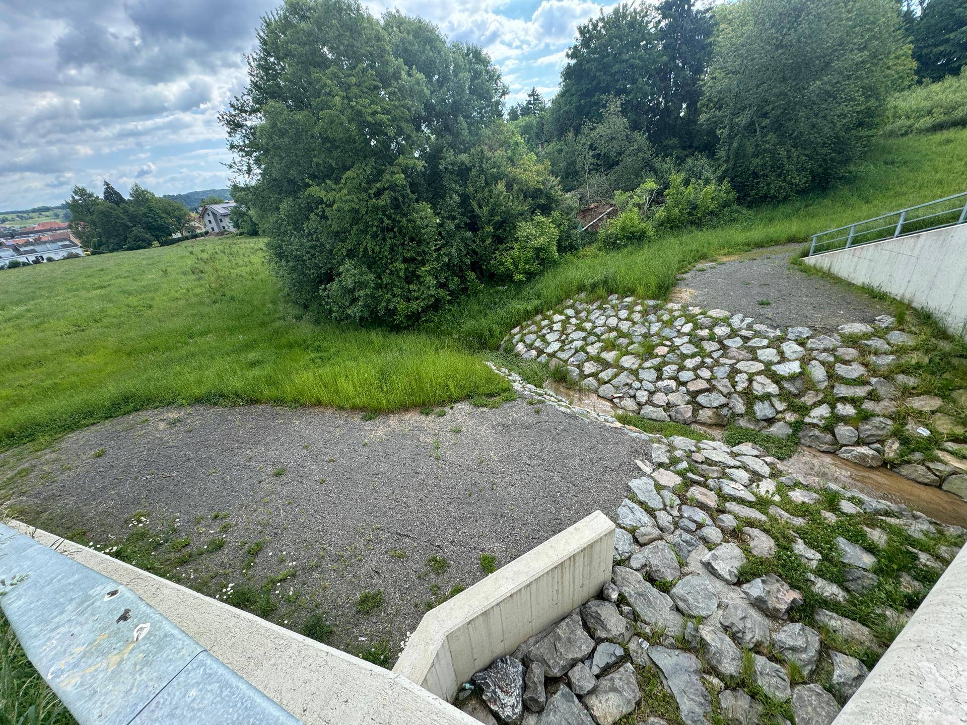 Betonierte Entwässerungsanlage mit Steinen und grünen Hängen in einer ländlichen Umgebung.
