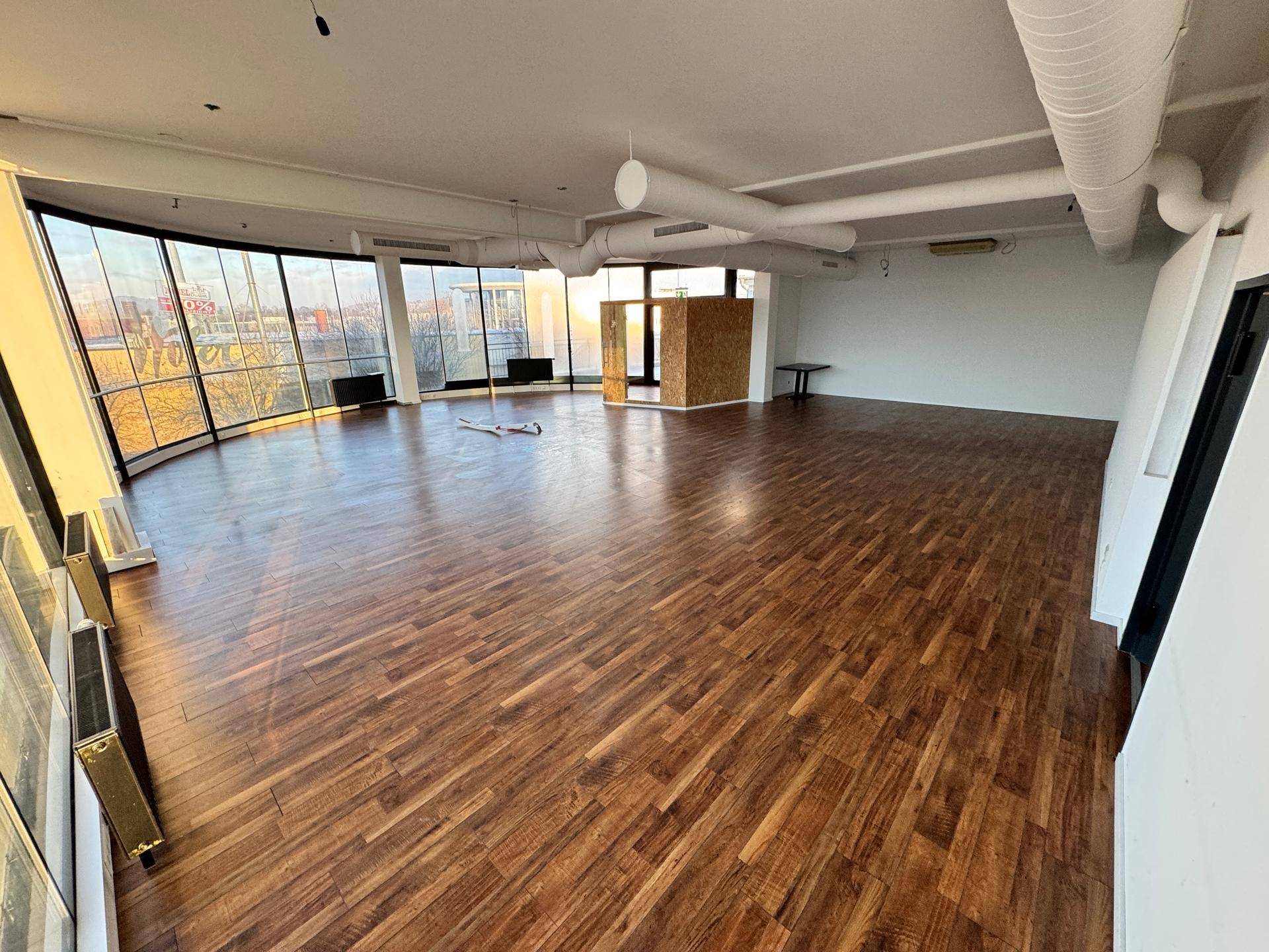 Weitläufiger Innenraum mit Holzboden, freiliegenden Rohren an der Decke und Panoramablick durch große Fenster.
