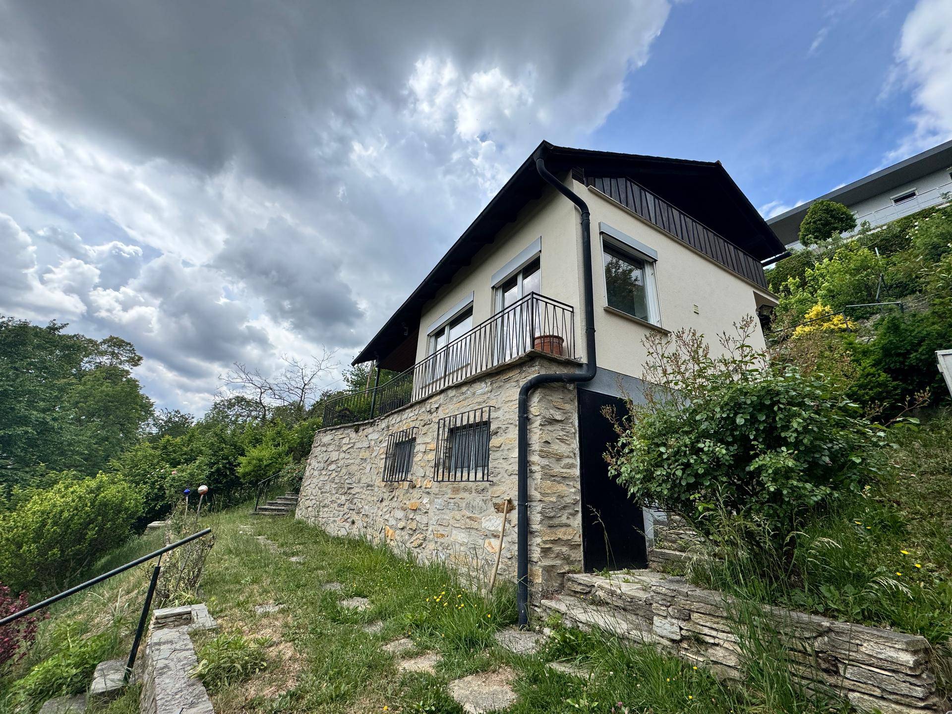 Hausfassade mit Steinsockel und Balkon, eingebettet in eine grüne Hanglage unter bewölktem Himmel.
