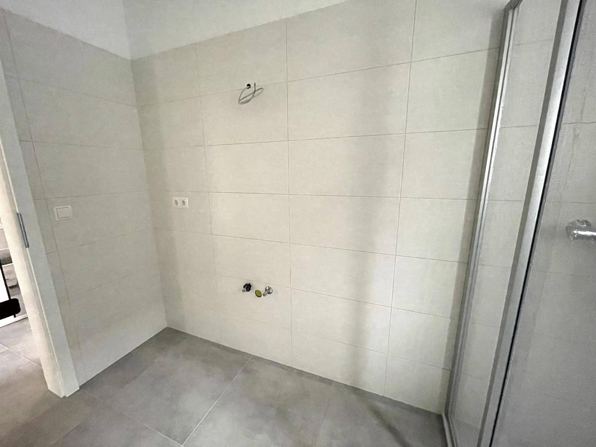 Modernes Badezimmer mit hellen Fliesen und Anschlüssen für Waschbecken und Waschmaschine.