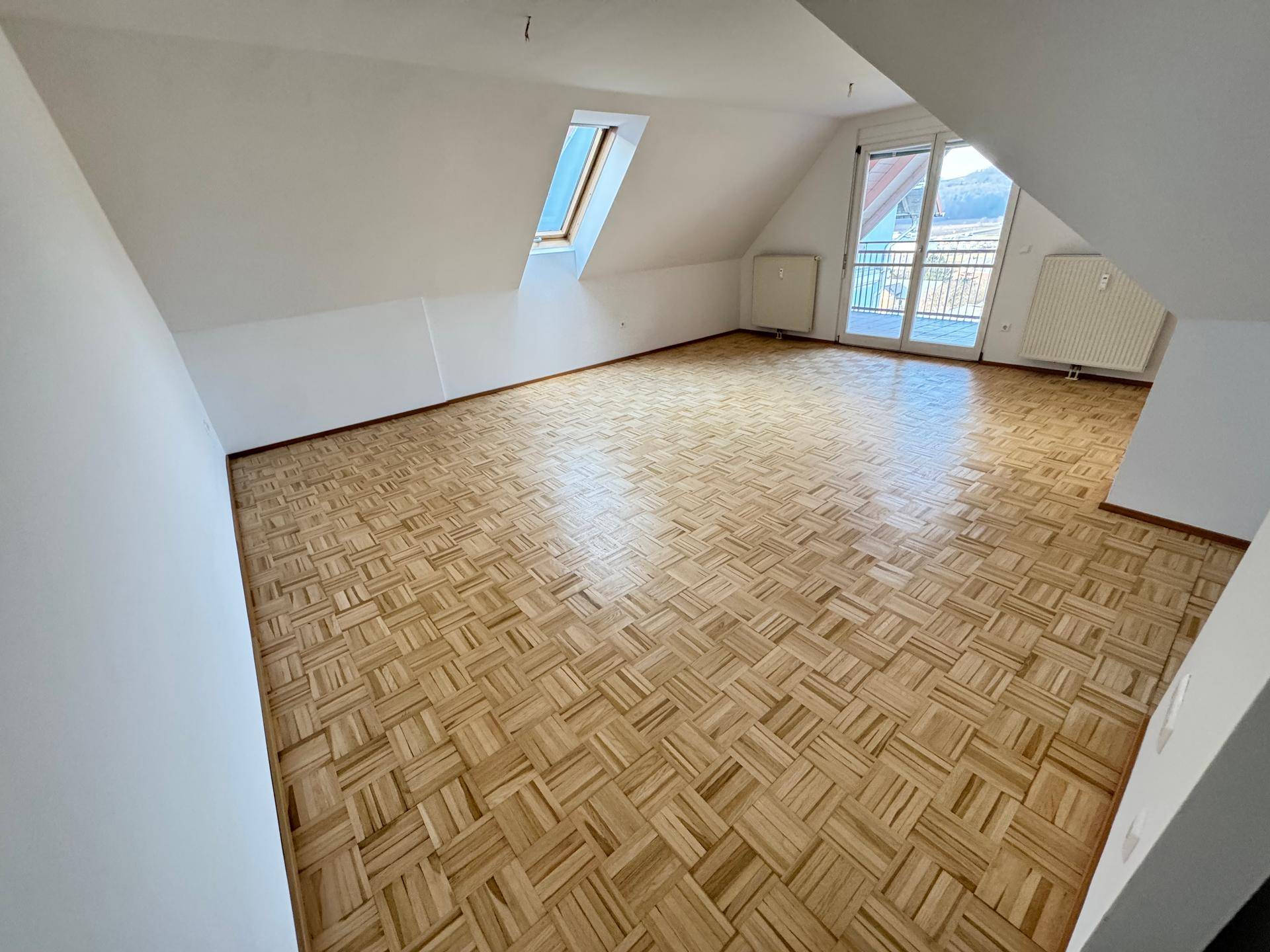 Großes Zimmer im Dachgeschoss mit Parkettboden, Dachschrägen, Dachfenster und Balkonzugang.