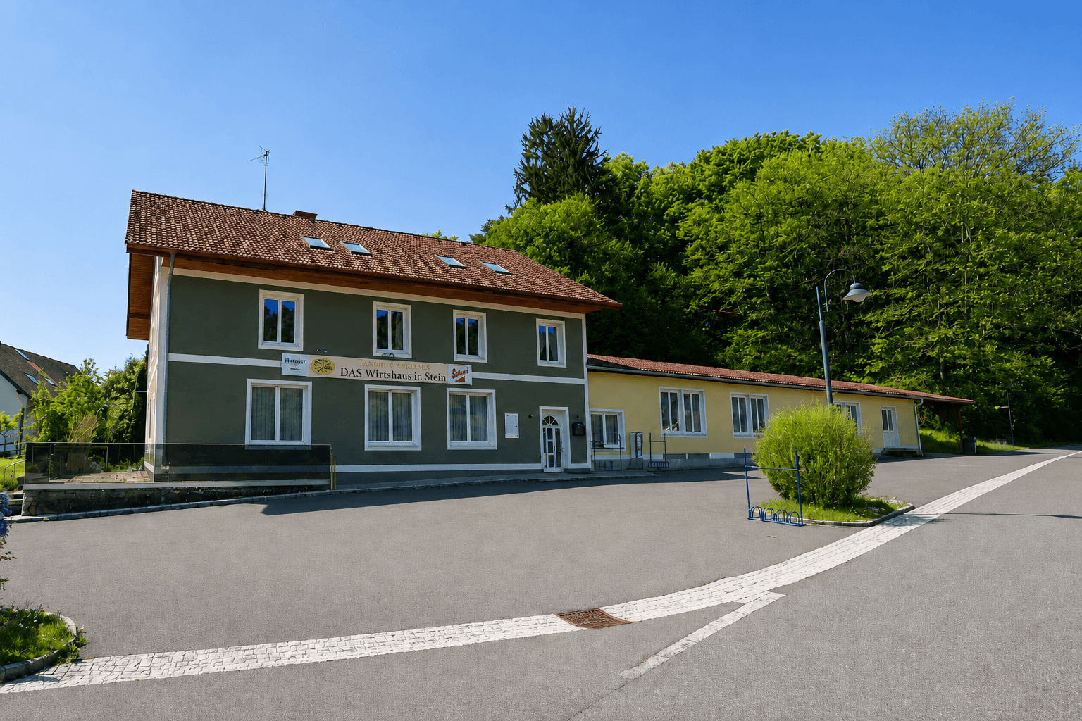 Ein traditionelles Gebäude mit grün-grauer Fassade und rotem Ziegeldach, umgeben von Bäumen und einem Parkplatz.