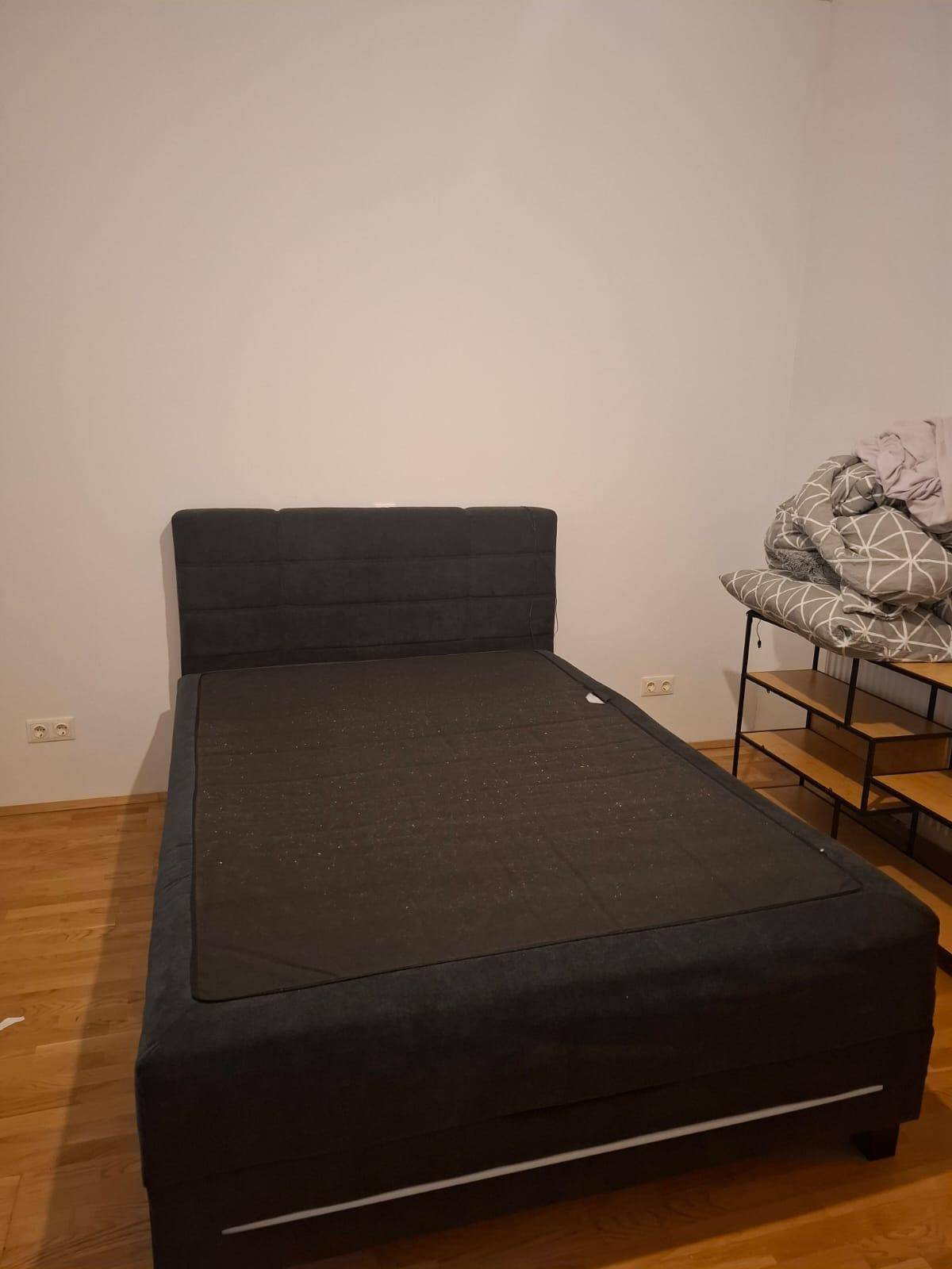 Schlafzimmer mit dunklem Bettrahmen, Holzboden und Ablageregal an der Seite.
