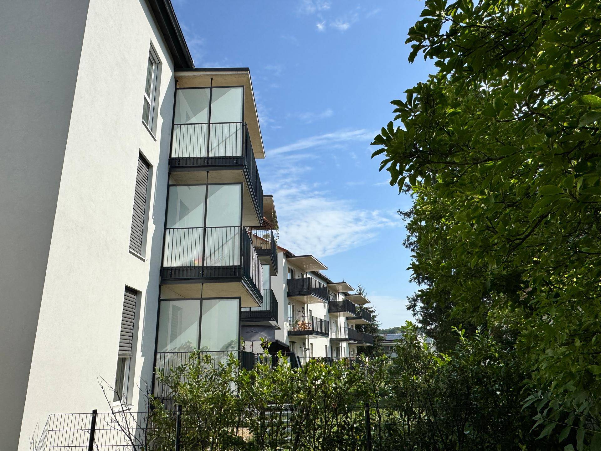 Moderne Wohnfassade mit Balkonen, umgeben von üppigem Grün und Bäumen unter blauem Himmel.