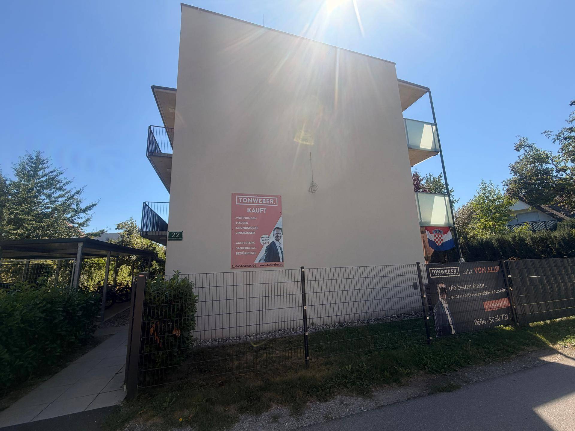 Seitenansicht des Gebäudes mit einer Immobilienwerbung und einem Zaun im Vordergrund.